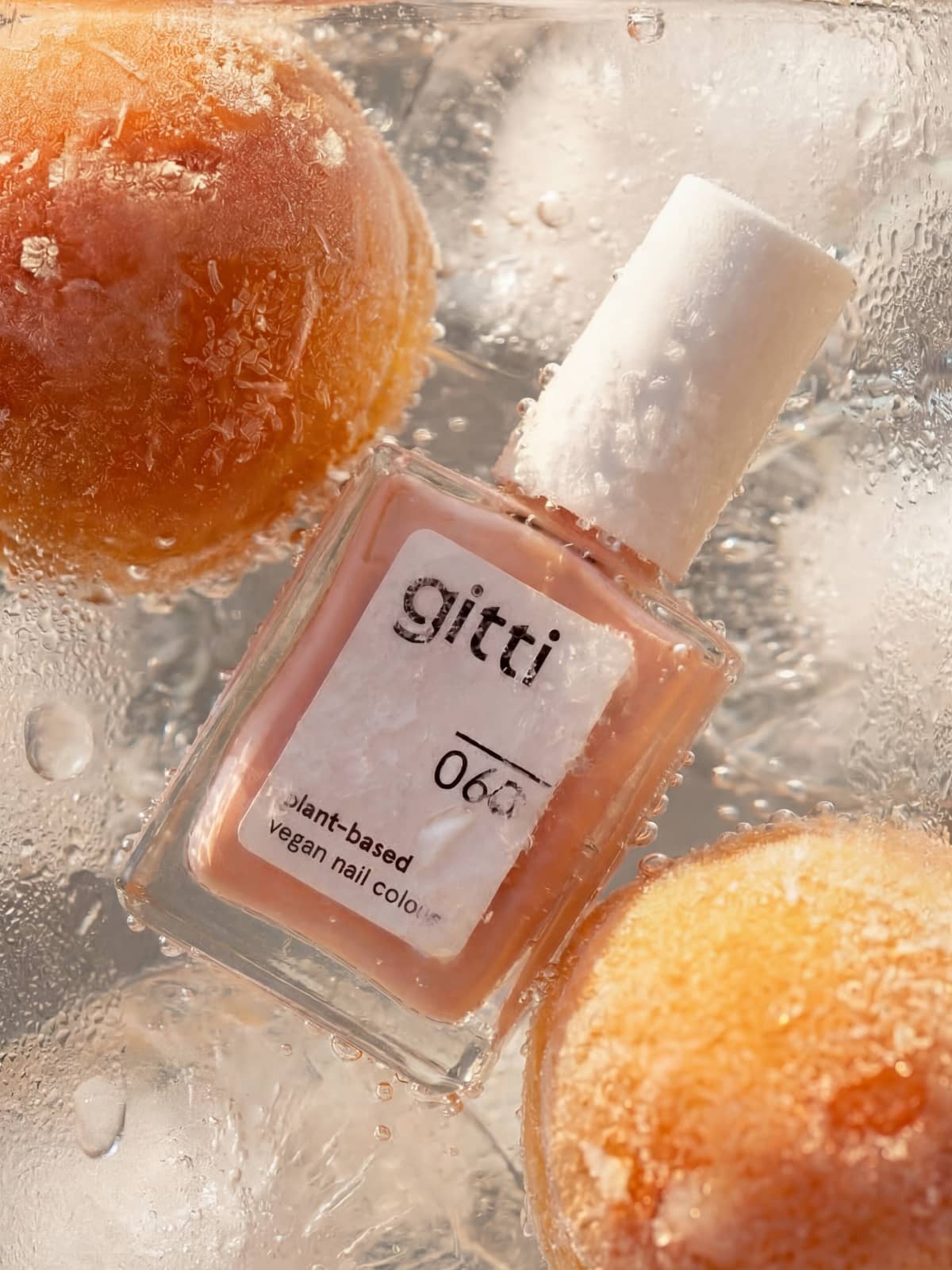 Eine Flasche des veganen Nagellacks „Gitti Nr. 060“ in der Farbe „Transparentes Apricotton“ steht auf Eis neben zwei gefrorenen Orangenkugeln und strahlt einen frischen Glanz aus, während sich Kondenswassertropfen am Glas bilden.
