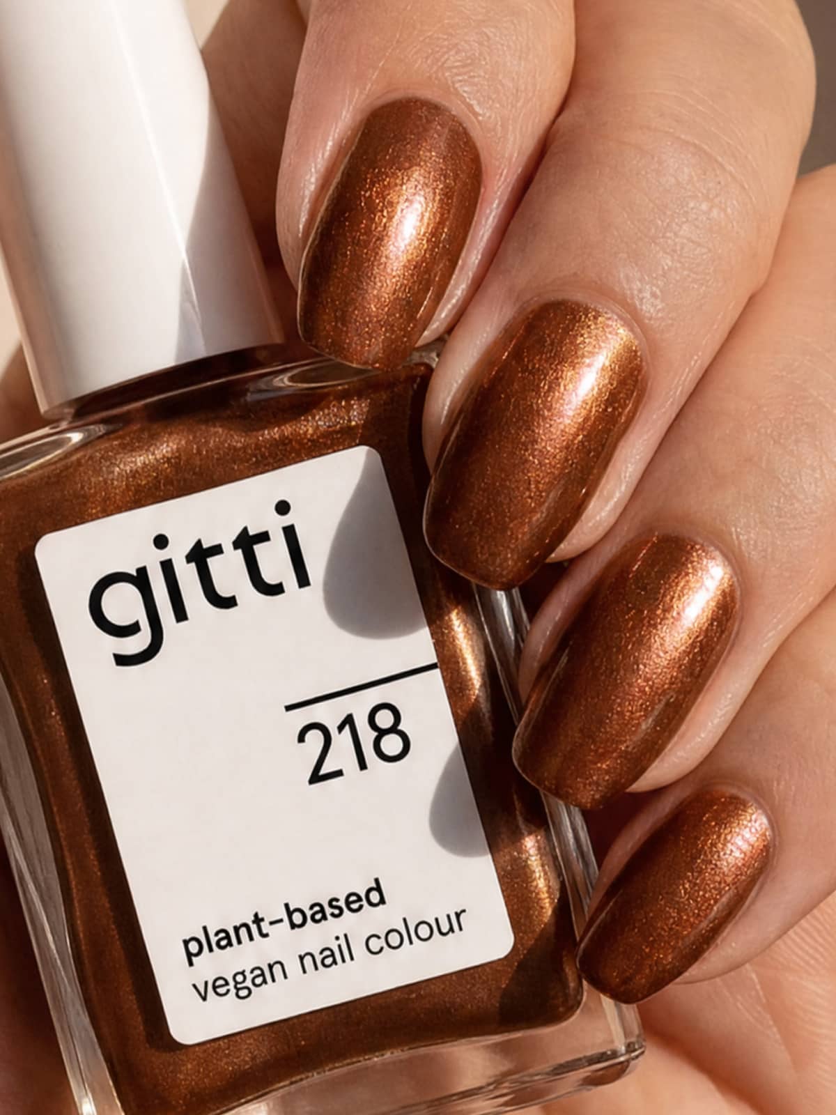 Eine Hand mit metallisch-bronzefarbener Nagellackierung hält eine Flasche aus dem „Colour & Concentrate Trio“ in der Hand, einer pflanzenbasierten, veganen Nagellackkollektion mit Farbtönen wie „gitti 218“, die pflanzenbasierte Farben präsentiert.