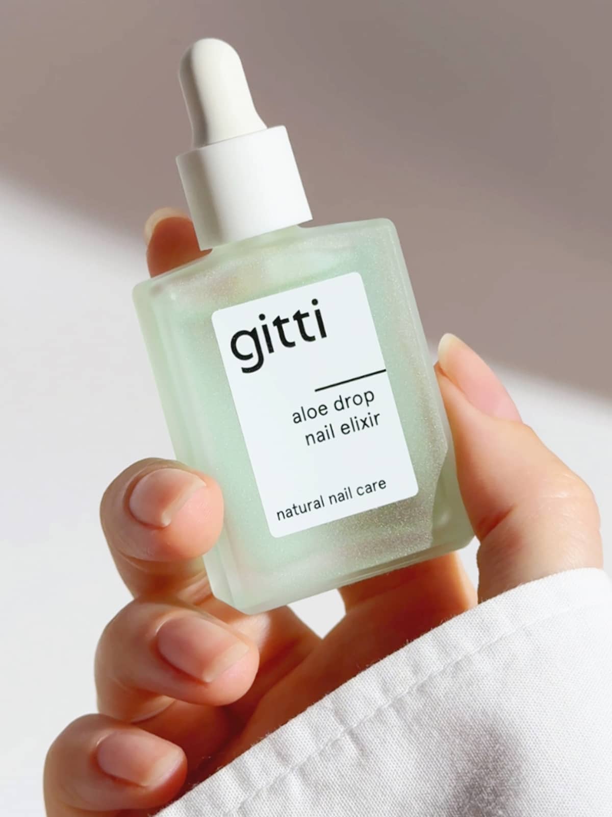 Vor einem neutralen Hintergrund ist eine Hand zu sehen, die eine Tropfflasche aus Milchglas mit der Aufschrift „Aloe Drop Nail Concentrate“ hält – ein Gel-Serum für die natürliche Nagelpflege und gesunde Nägel.