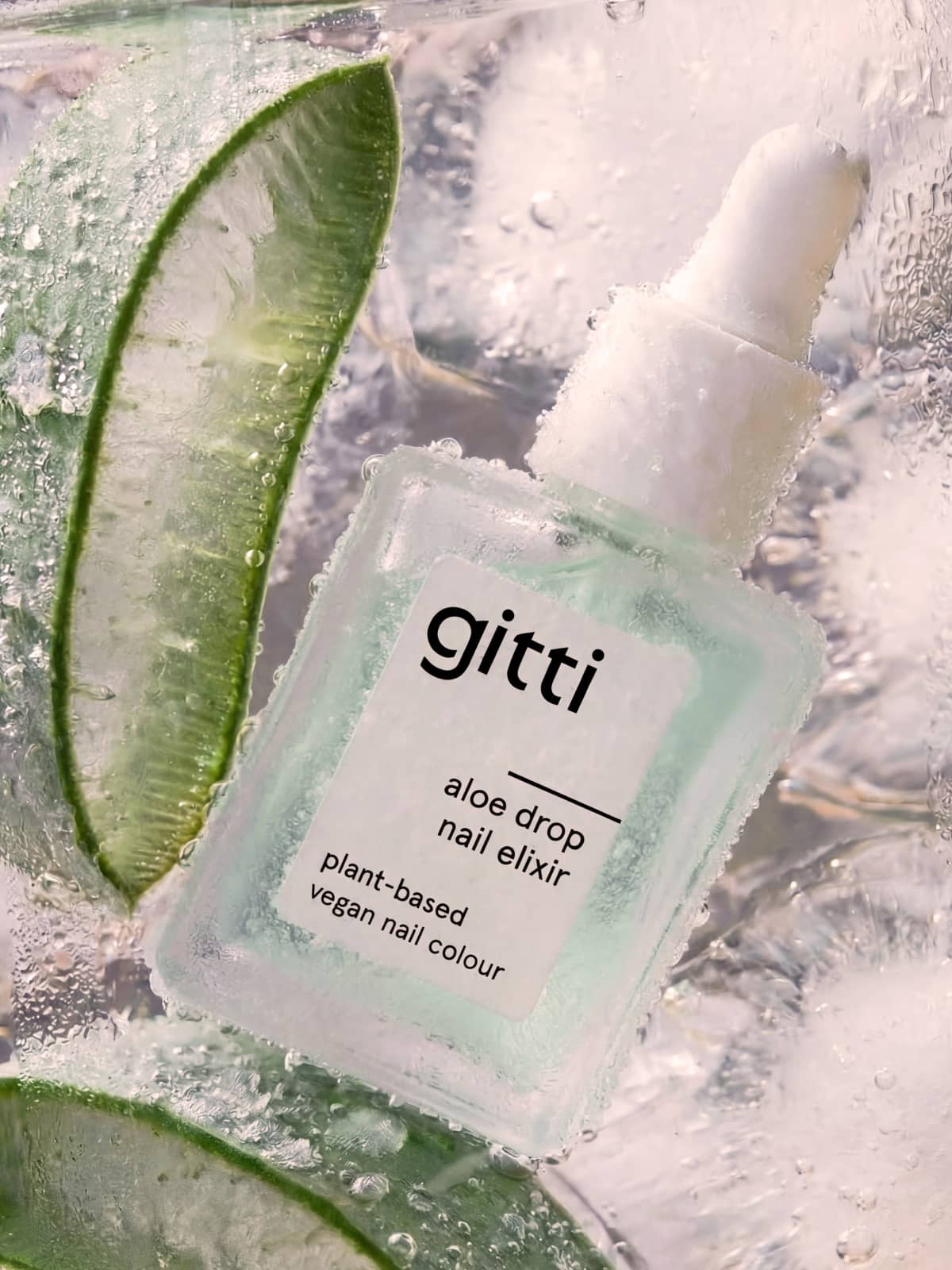 Eine elegante Tropfflasche aus Milchglas mit der Aufschrift „gitti Aloe Drop Nail Concentrate“ steht auf Eis neben frischen Aloe-Scheiben. Dieses belebende Gel-Serum pflegt sowohl die Nagelhaut als auch die Nägel. Der Text lautet: pflanzliche, vegane Nagelpflege.