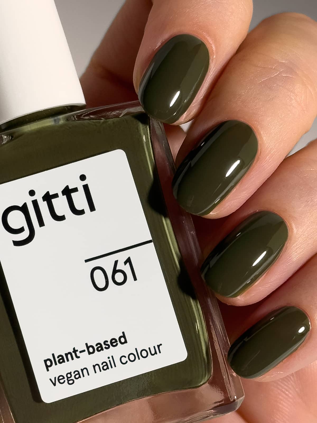 Eine Hand mit dunkelolivgrün lackierten Fingernägeln hält eine Flasche „Gitti Nr. 061“, einen pflanzlichen, veganen Nagellack in einem passenden Farbton.