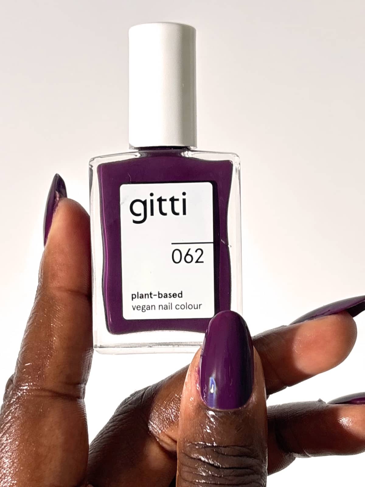 Eine Hand mit violett lackierten Nägeln hält vor einem schlichten weißen Hintergrund eine Flasche „Gitti Nr. 062“ in der Hand, einen pflanzlichen, veganen Nagellack in der Farbe „Pflaumenlila“ mit einem rötlichen Unterton.