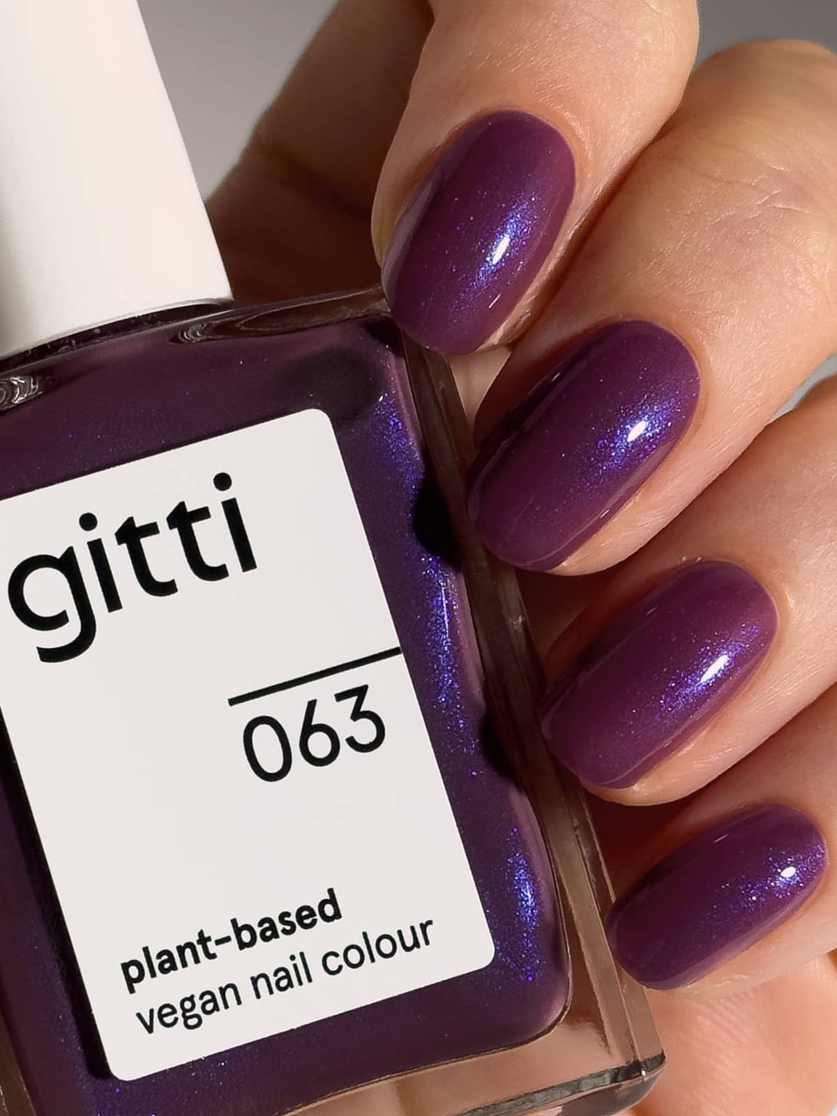 Eine Hand mit Nägeln, die in einem halbtransparenten Pflaumenton lackiert sind, hält eine Flasche „Gitti Nr. 063“, einen pflanzlichen, veganen Nagellack.