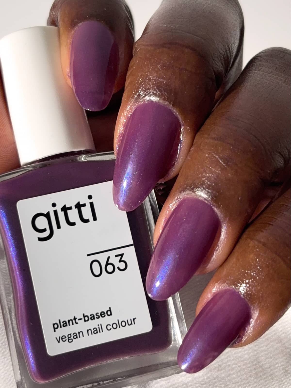 Eine Hand mit langen Nägeln, die in einem halbtransparenten Pflaumenton lackiert sind, hält eine Flasche „Gitti Nr. 063“ – einen pflanzlichen, veganen Nagellack in einem passenden Farbton.