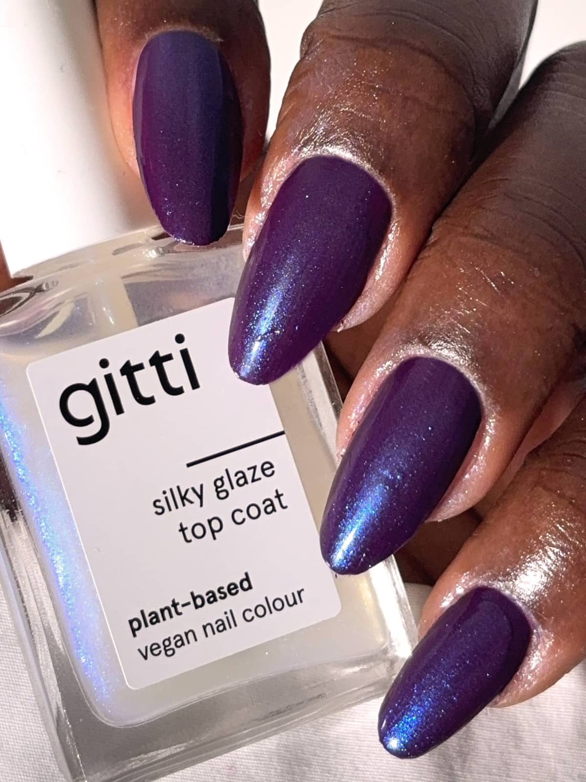 Eine Hand mit dunkelviolettem, schimmerndem Nagellack, der einen Hauch von blauem Schimmer aufweist, hält eine quadratische Flasche mit der Aufschrift „Silky Glaze Top Coat“ – ein pflanzlicher, veganer Nagellack, der für ein strahlendes, glasiges Finish sorgt.