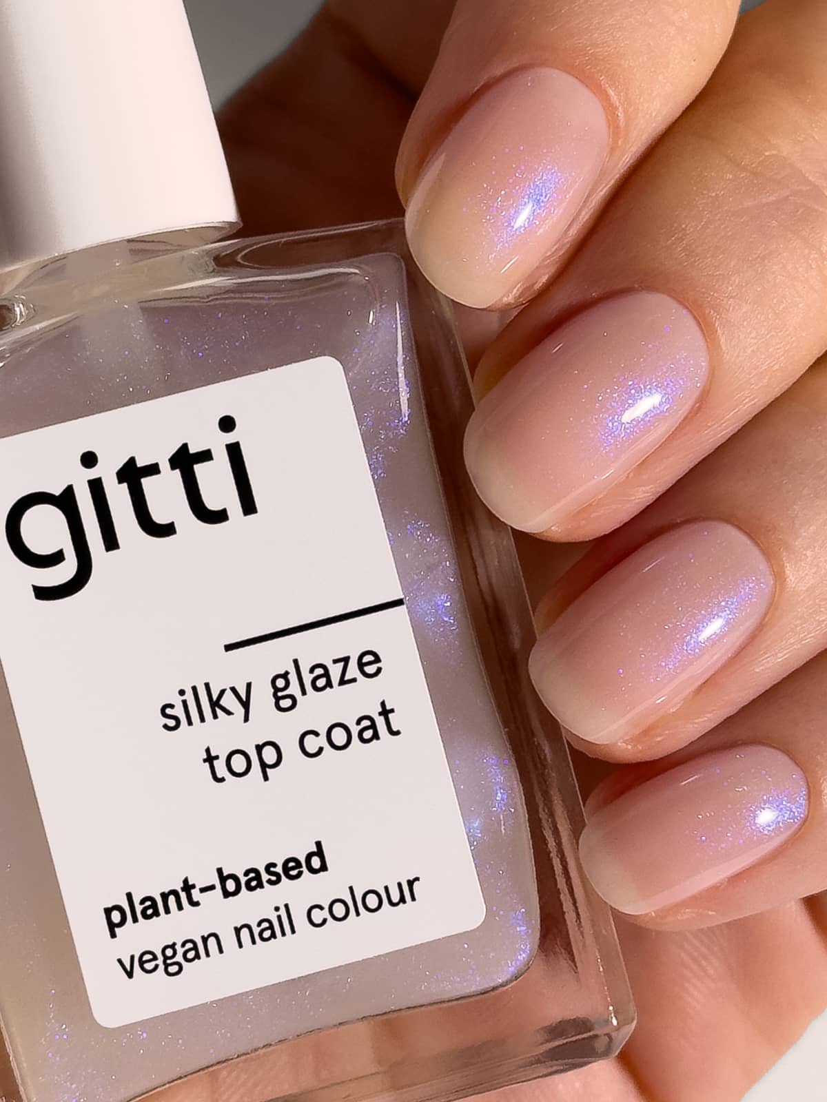 Eine Hand mit kurzen, ovalen Nägeln, die mit einem transparenten, schillernden Nagellack mit dezentem blauem Schimmer lackiert sind, hält eine Flasche mit der Aufschrift „Silky Glaze Top Coat“ – ein pflanzlicher, veganer Nagellack für ein perfektes, glasiertes Finish.