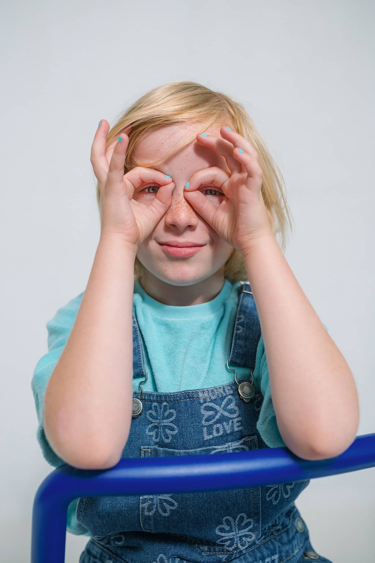 Ein lächelndes blondes Kind in blauer Latzhose und türkisem Hemd bastelt mit seinen Händen Gläser und präsentiert fröhlich die gitti kids no. 01-sichere Kinderkosmetik für die Kleinsten.