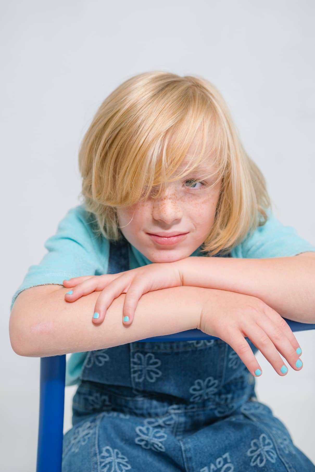 Ein Kind mit langen blonden Haaren und hellblauem Gitti Kids No. 01 Nagellack lehnt sich mit verschränkten Armen auf einem blauen Stuhl nach vorne und trägt ein türkisfarbenes Hemd und einen Jeans-Overall mit einem Blumenmuster.