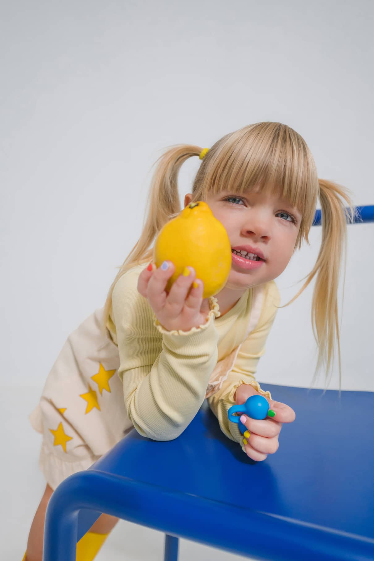Ein junges Mädchen mit blonden Zöpfen lehnt auf einem blauen Stuhl, hält eine Zitrone und ein Spielzeug in der Hand und trägt ein helles Outfit mit gelben Sternen. Stolz zeigt sie gitti kids no. 06, ihren bunten, ungeschenkten Nagellack, auf ihren winzigen Fingernägeln.