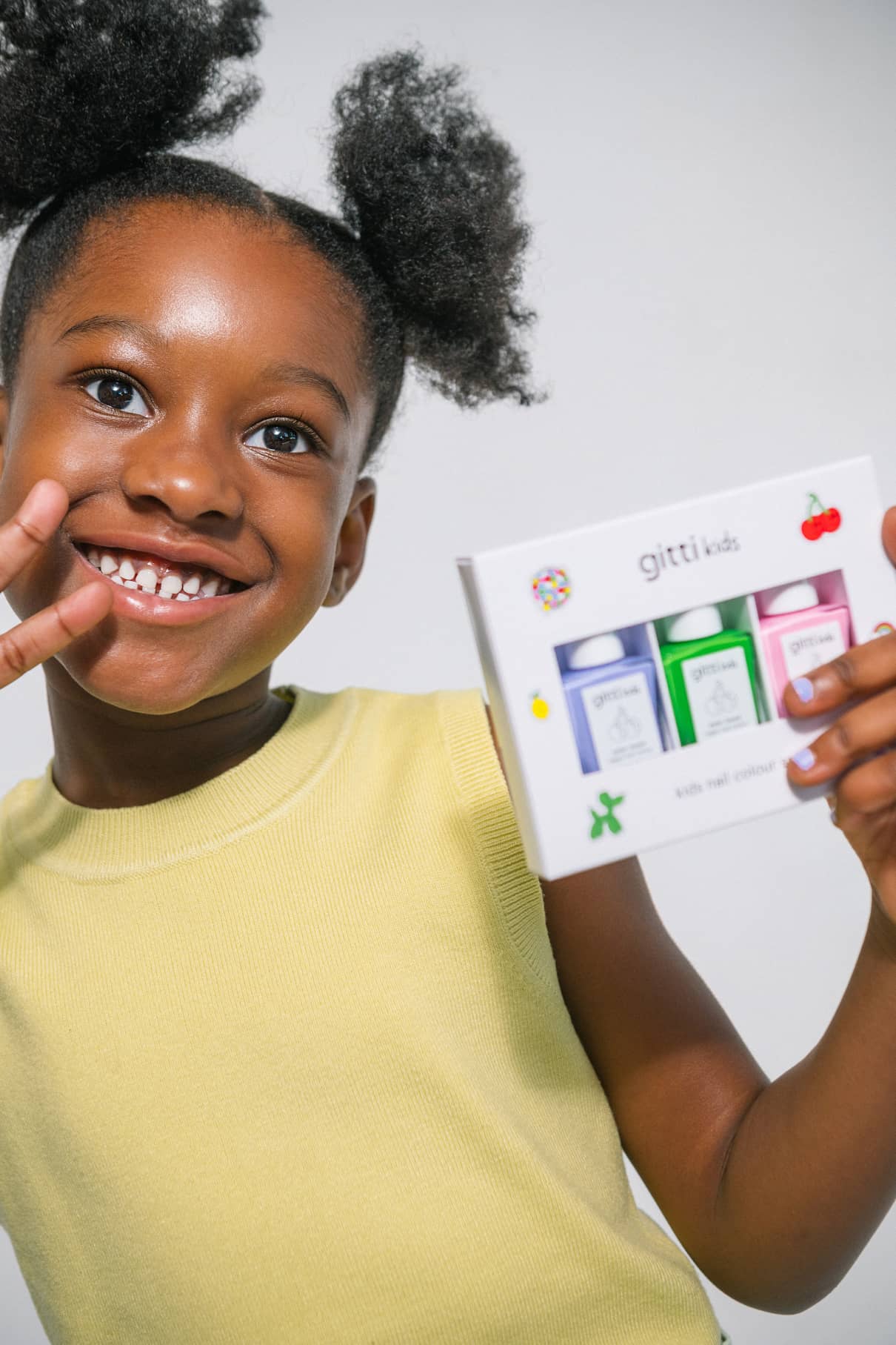 Ein lächelndes Kind in einem gelben ärmellosen Hemd hält eine Schachtel mit dem Little Artist Set hoch und zeigt mit der anderen Hand ein Friedenszeichen.