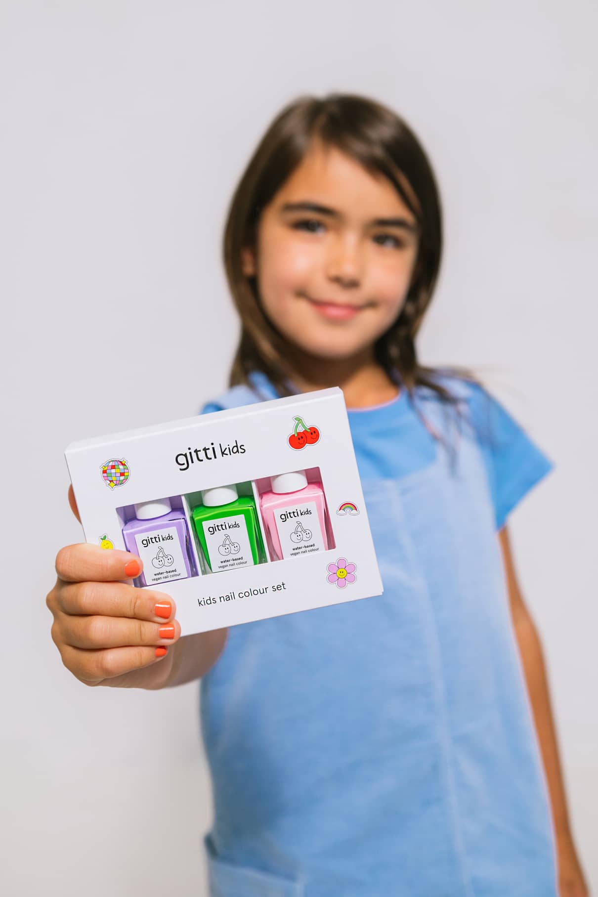 Ein Mädchen in einem blauen Hemd hält das Little Artist Set mit den Farbtönen La La Lilac und Green Planet in der Hand und präsentiert drei Nagellackflaschen vor einem schlichten weißen Hintergrund.