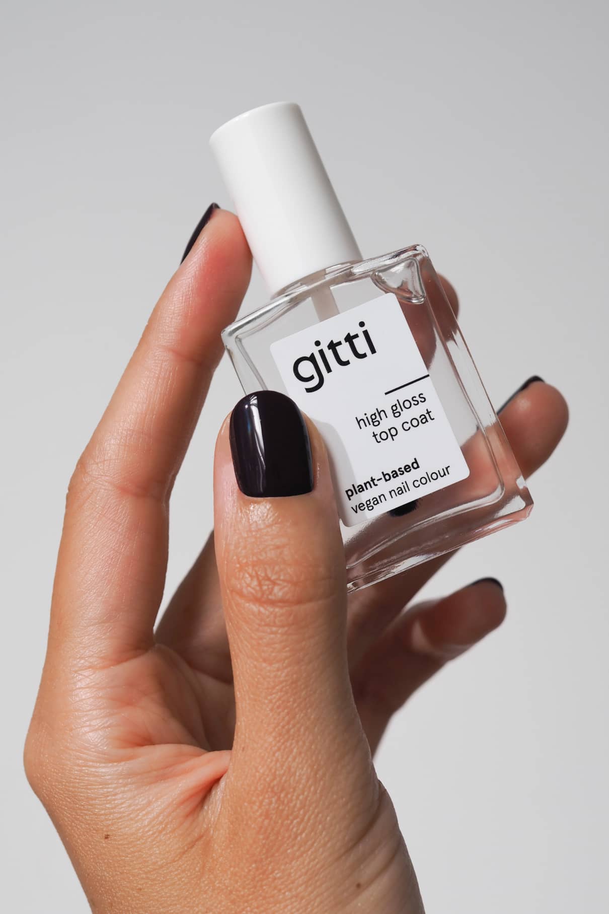Eine Hand mit dunkelviolettem Nagellack hält eine Flasche High Gloss Top Coat, der als pflanzliche und vegane Nagelfarbe für ein intensives Hochglanzfinish gekennzeichnet ist.