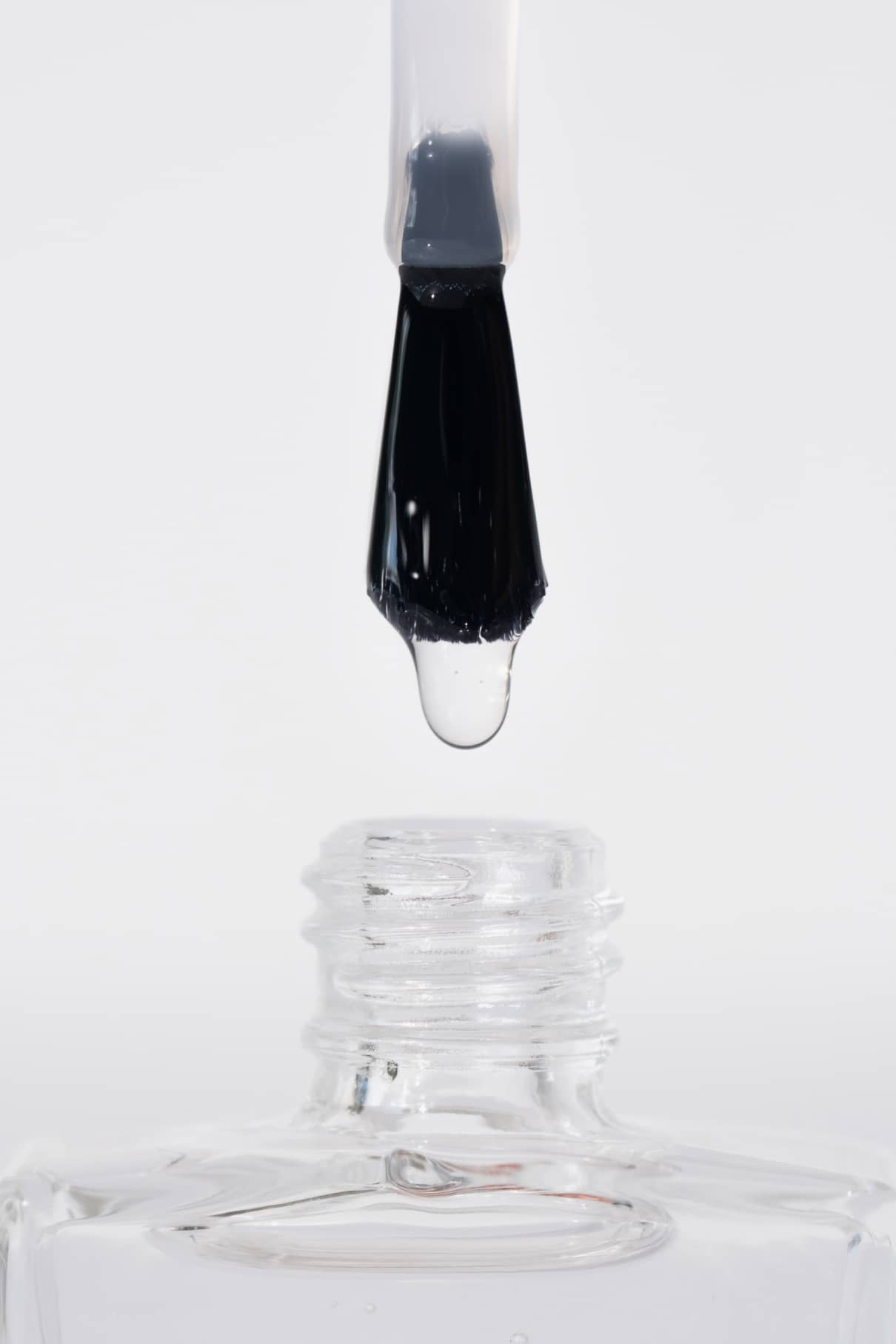Eine Nahaufnahme zeigt den Hochglanz-Topcoat-Pinsel mit einem klaren Tropfen, der über die offene Mündung der transparenten Flasche gehalten wird, vor einem weißen Hintergrund, der das brillante Hochglanz-Finish zur Geltung bringt.