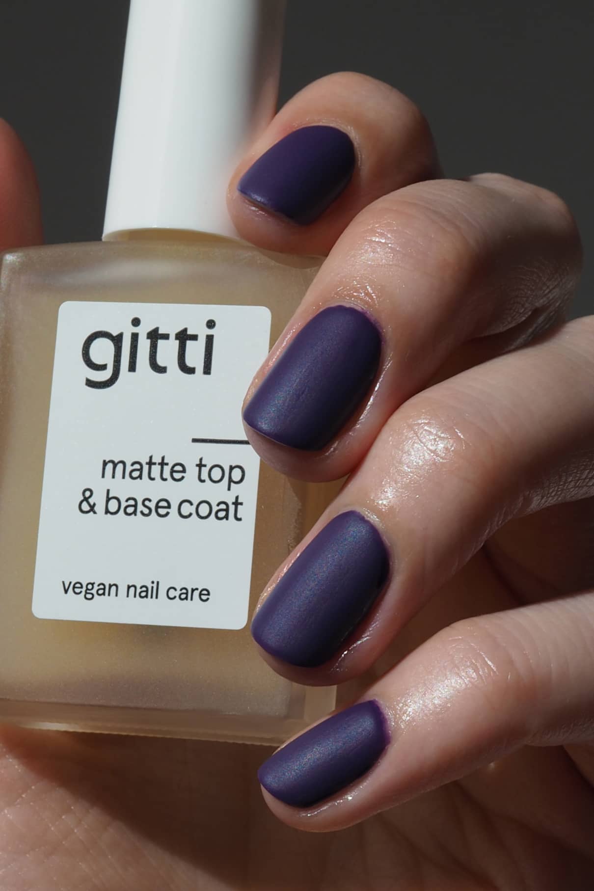 Eine Hand mit matten, dunkelvioletten Nägeln hält eine Flasche Matte Top & Base Coat - vegane Nagelpflege, die zusätzlichen Halt und ein mattes Finish in einem 2-in-1-Produkt bietet.