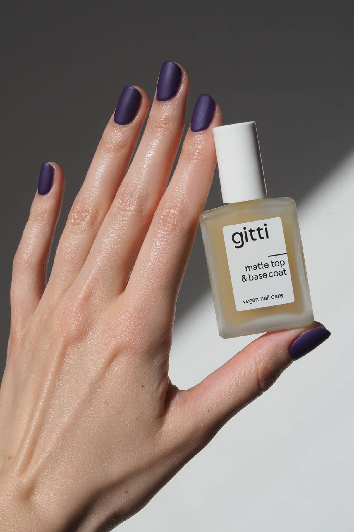 Eine Hand mit matten dunkelvioletten Nägeln hält eine Flasche Matte Top & Base Coat Nagelpflege für zusätzlichen Halt und ein mattes Finish vor einem grauen Hintergrund.