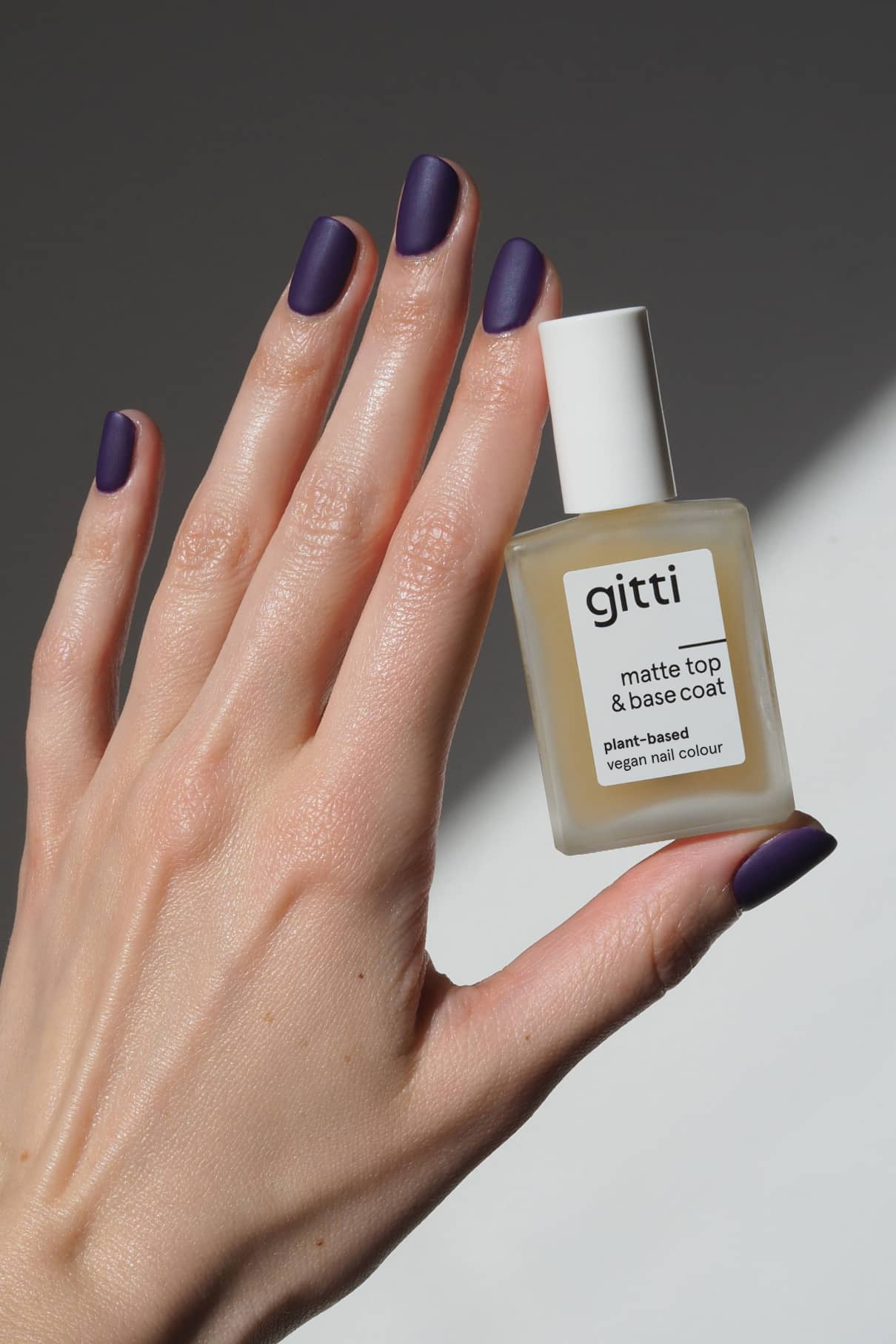 Eine Hand mit matten, dunkelvioletten Nägeln hält eine Flasche Matte Top & Base Coat, einen veganen Nagellack auf pflanzlicher Basis für zusätzlichen Halt und ein makelloses, mattes Finish, vor einem neutralen Hintergrund.