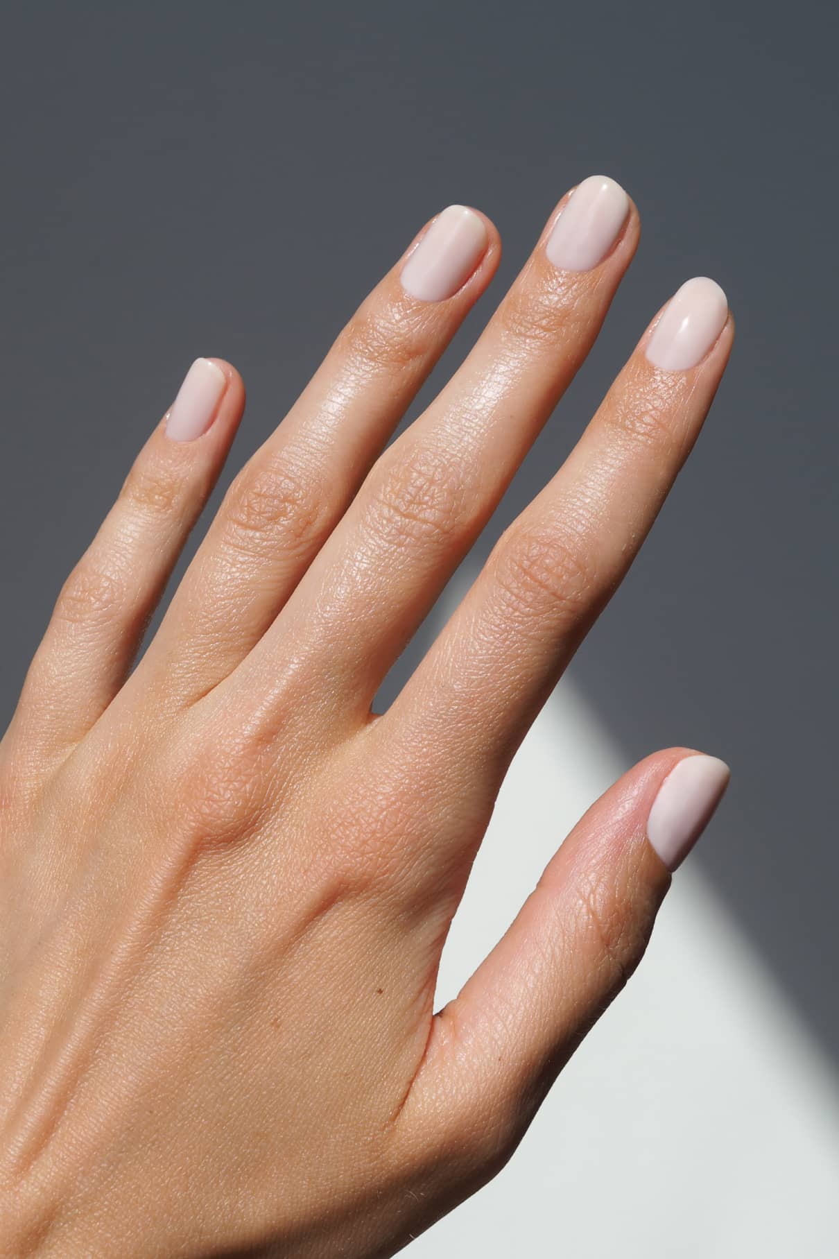 Eine Hand mit sauber manikürten Nägeln, die mit Nail Glaze, einem halbtransparenten, milchigen Weiß, poliert sind, wird vor einem neutralen Hintergrund gezeigt.