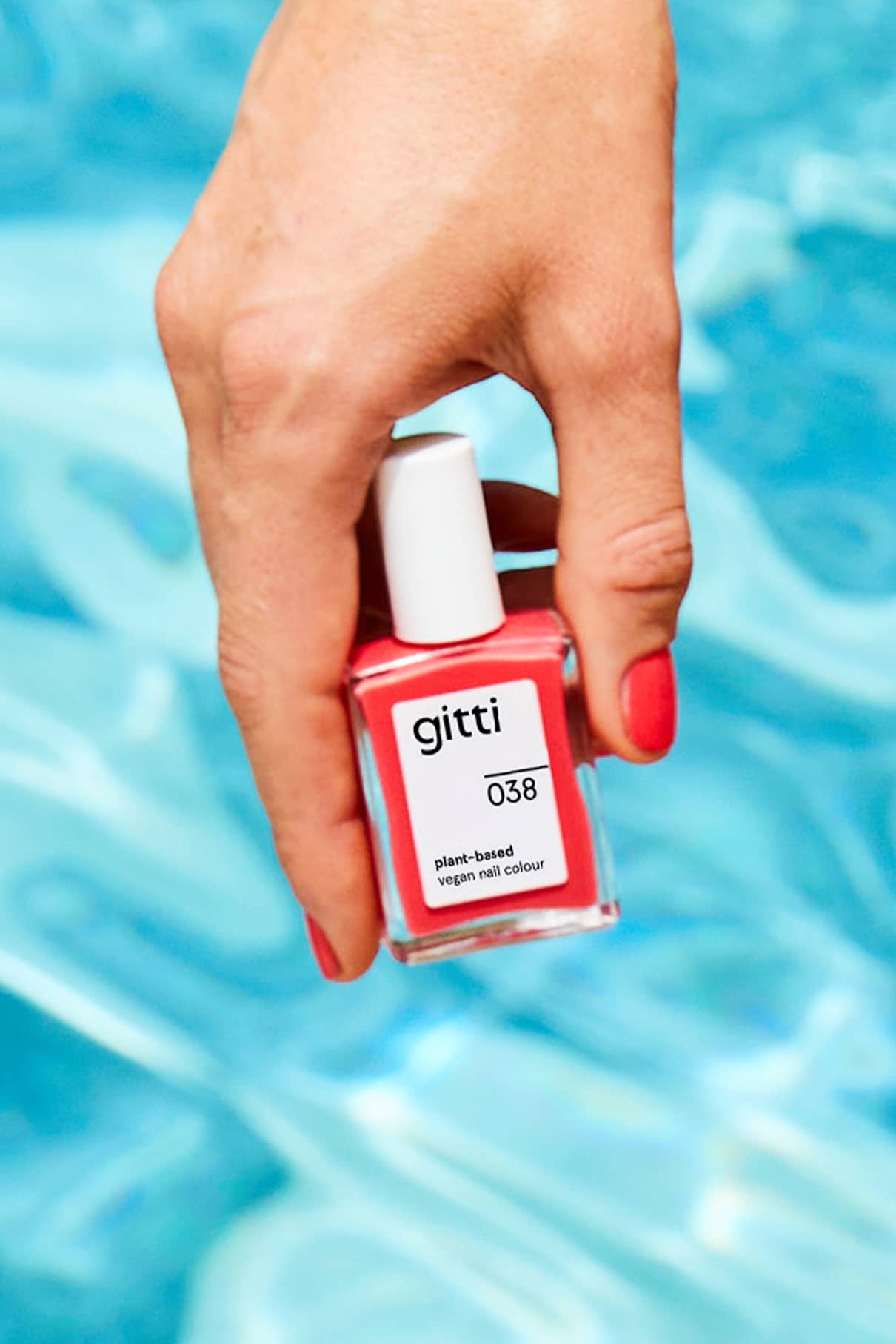 Eine Hand mit lackierten Nägeln hält ein Fläschchen mit gitti no. 038 pflanzliche vegane Nagelfarbe in Korallrot, die vor blauem Hintergrund Wärme und Tiefe ausstrahlt.