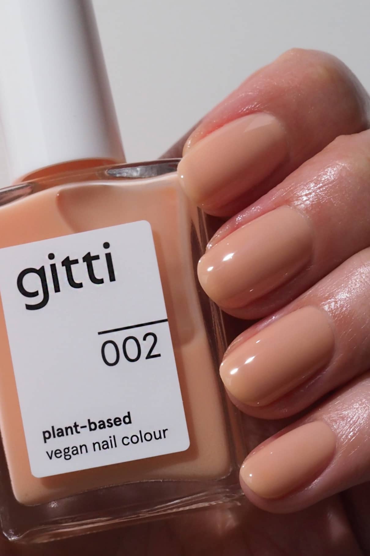 Eine Hand mit kurzen, sauber manikürten Nägeln, die in einem Pfirsichton lackiert sind, der von Pantones Farbe des Jahres 2024 inspiriert ist, hält eine Flasche gitti no. 002, einer veganen Nagelfarbe auf Pflanzenbasis.