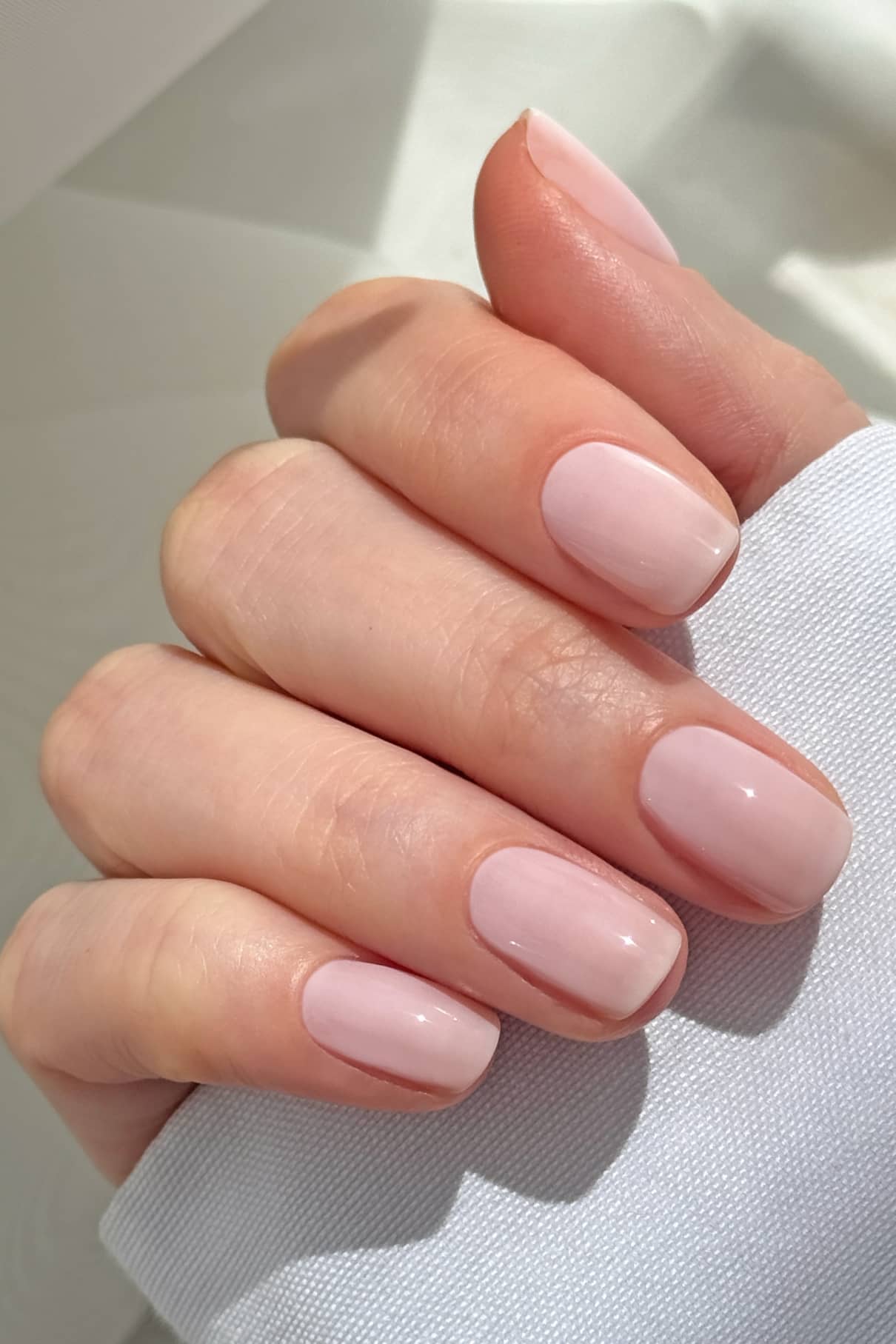 Eine Hand mit kurzen, sauber manikürten Nägeln, die mit dem Mini Getaway Set in einem glänzenden, blassrosa Farbton lackiert wurden, ruht auf einer leichten Stoffoberfläche.
