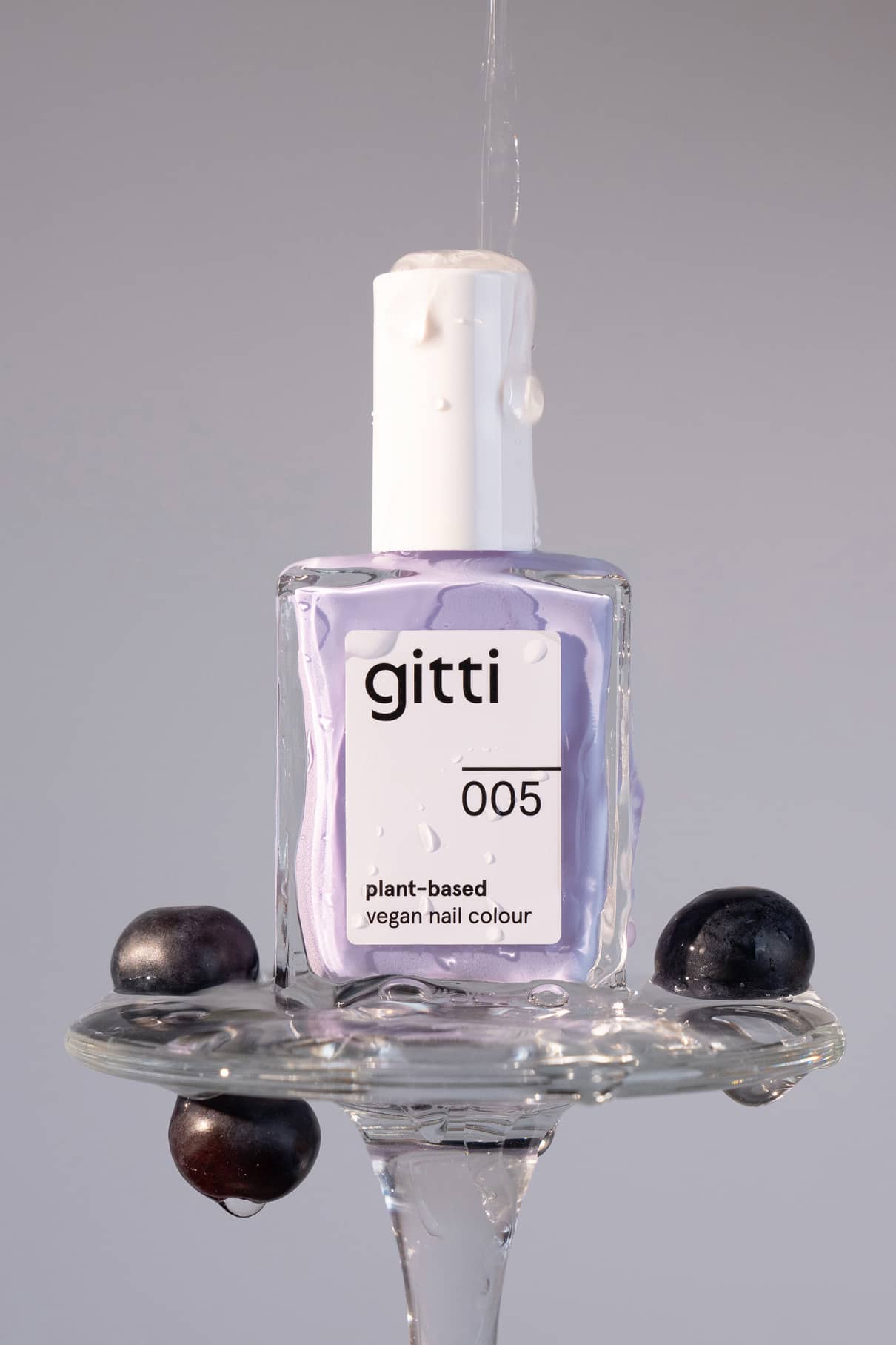 Eine Flasche gitti no. 005 pflanzlicher veganer Nagellack in harmonischem Lila ruht auf einer Glasfläche mit Wassertropfen und zwei schwarzen Weintrauben.