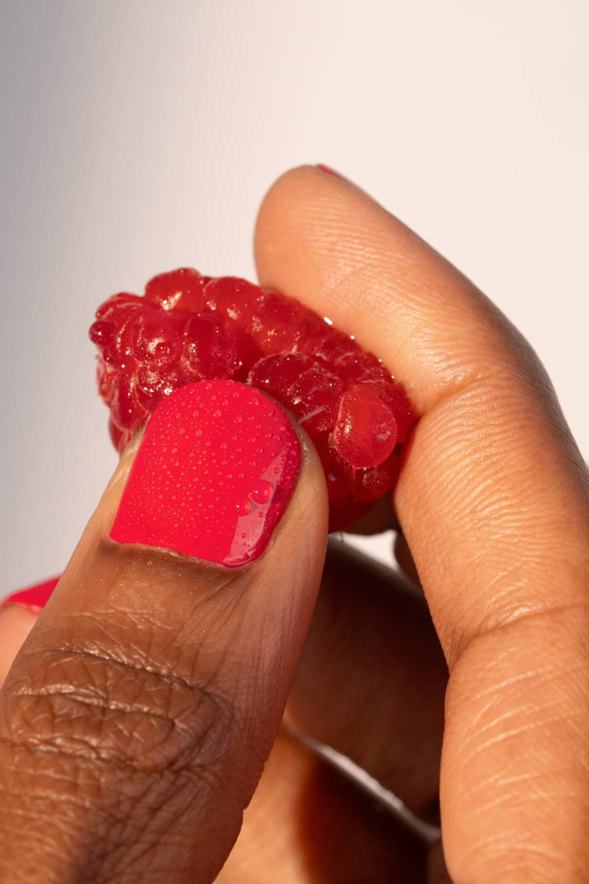 Eine Hand mit glänzendem gitti no. 006 Nagellack, einem leuchtenden Rot mit rosa Unterton, hält eine frische Himbeere zwischen Daumen und Zeigefinger vor einem schlichten Hintergrund.