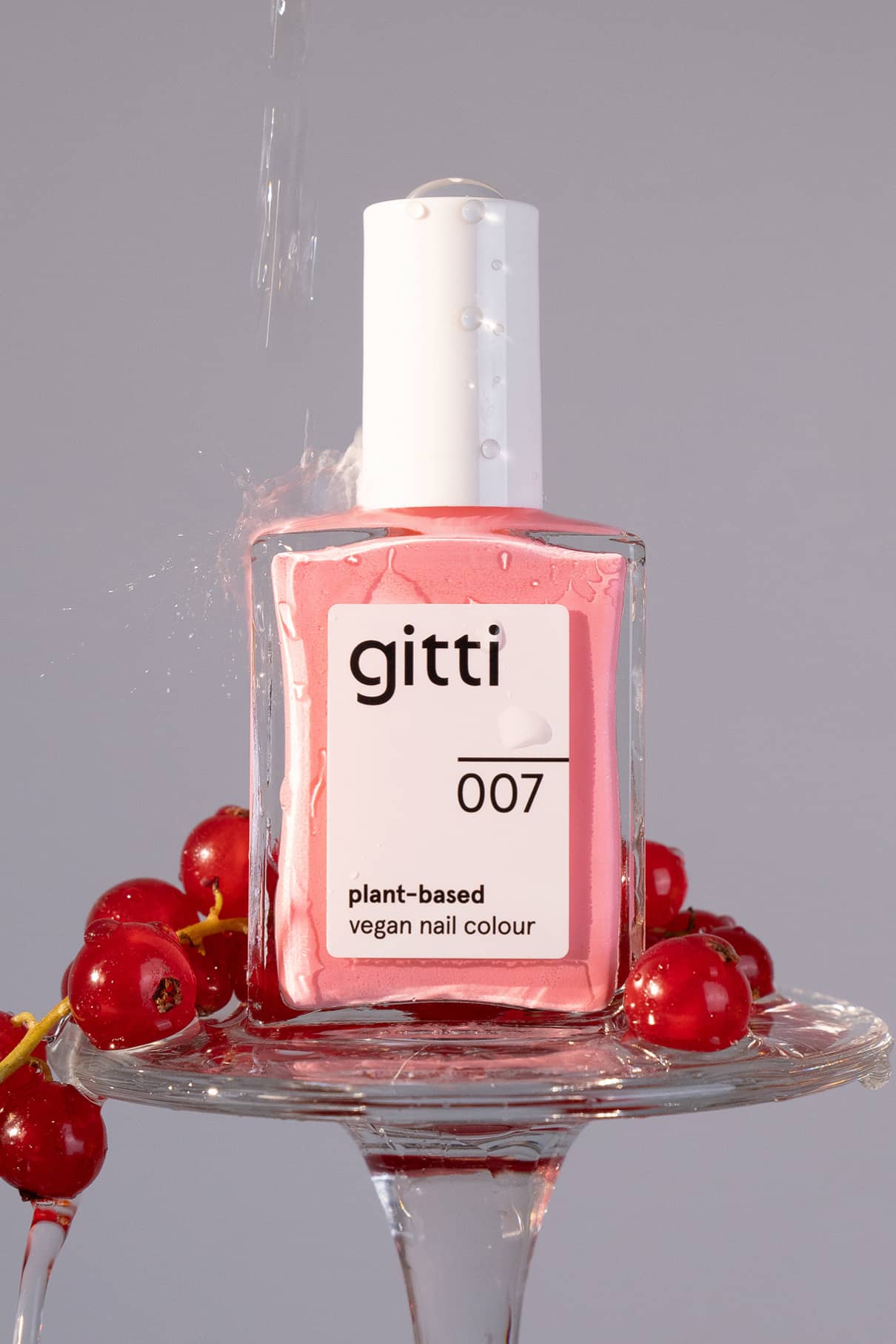 Eine Flasche gitti no. 007 pflanzlicher veganer Nagellack in leuchtendem Pink ruht auf einem Glasteller mit roten Johannisbeeren, über den Wasser spritzt.