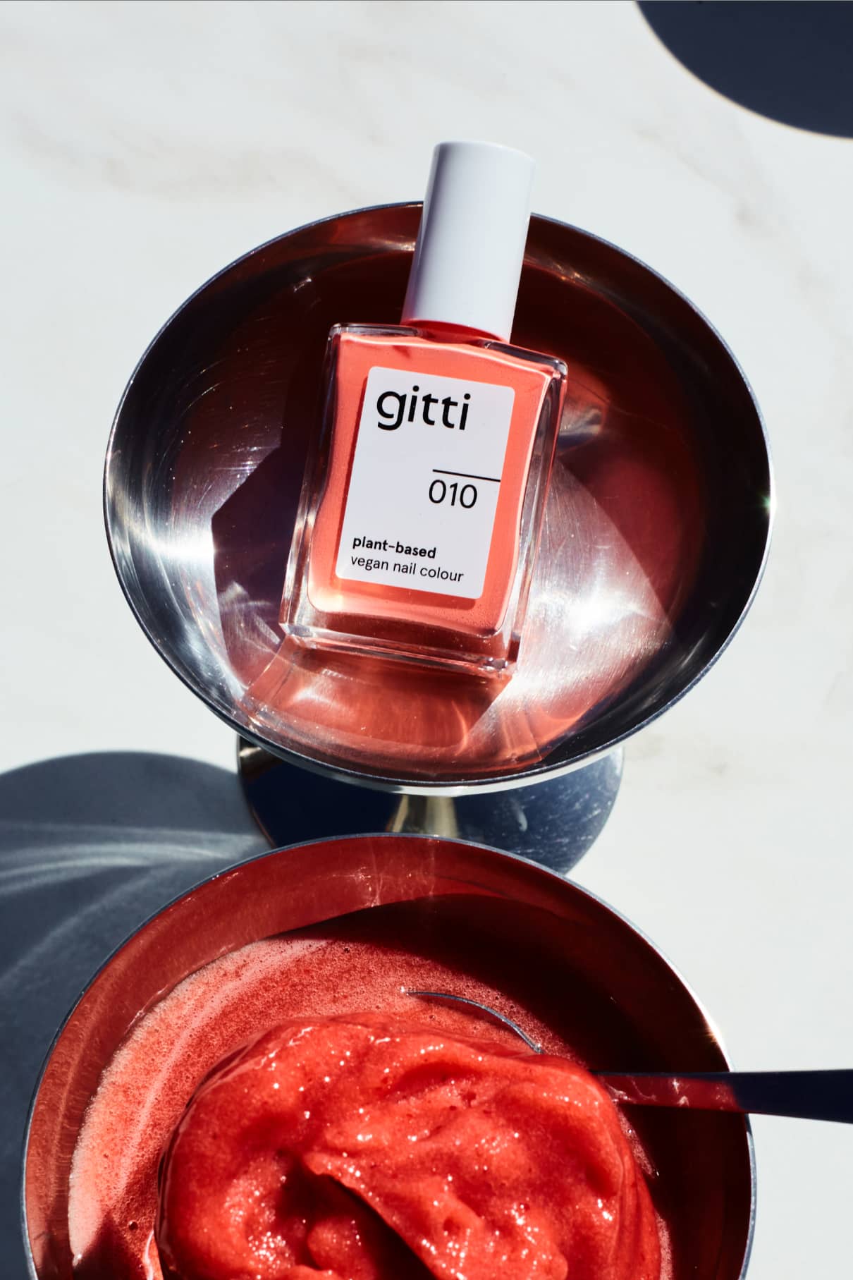 Eine Flasche gitti no. 010, einer veganen Nagelfarbe auf Pflanzenbasis, die für ihren intensiven Korallton bekannt ist, steht in einer Metallschale neben einer Schale mit roter, schaumiger Flüssigkeit und einem Löffel.