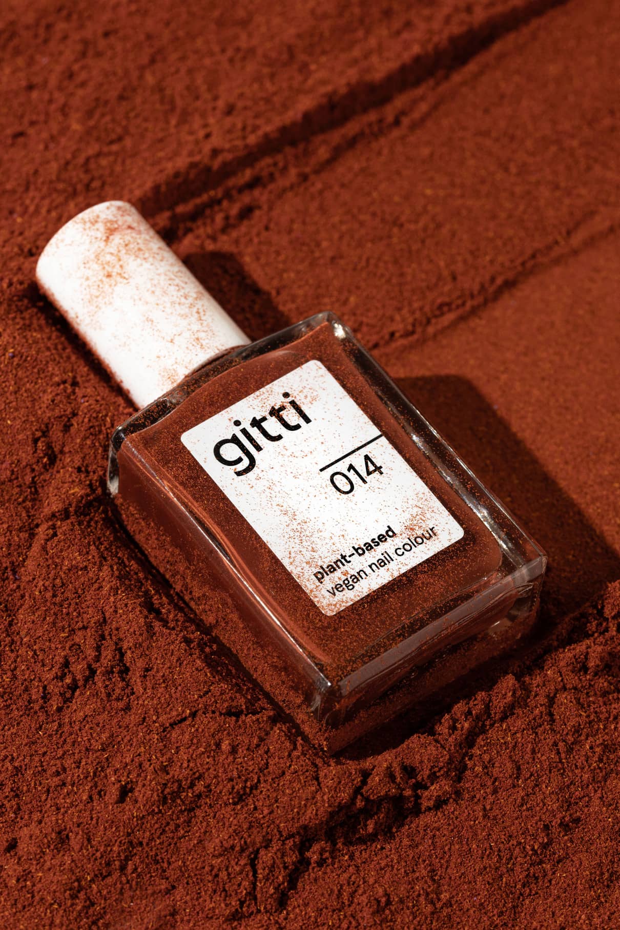 Eine Flasche mit gitti no. 014 pflanzliche, vegane Nagelfarbe ruht auf einer mit warmem, rötlich-braunem Puder bestäubten Oberfläche.