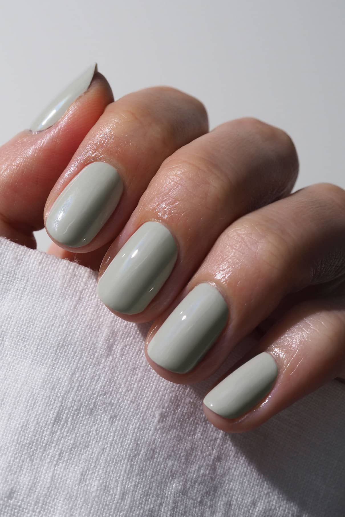 Eine Hand mit gepflegten Nägeln in gitti no. 017, einem hellen Grau mit salbeigrünem Unterton, hält einen weißen Stoff - die winterliche Version moderner Eleganz.