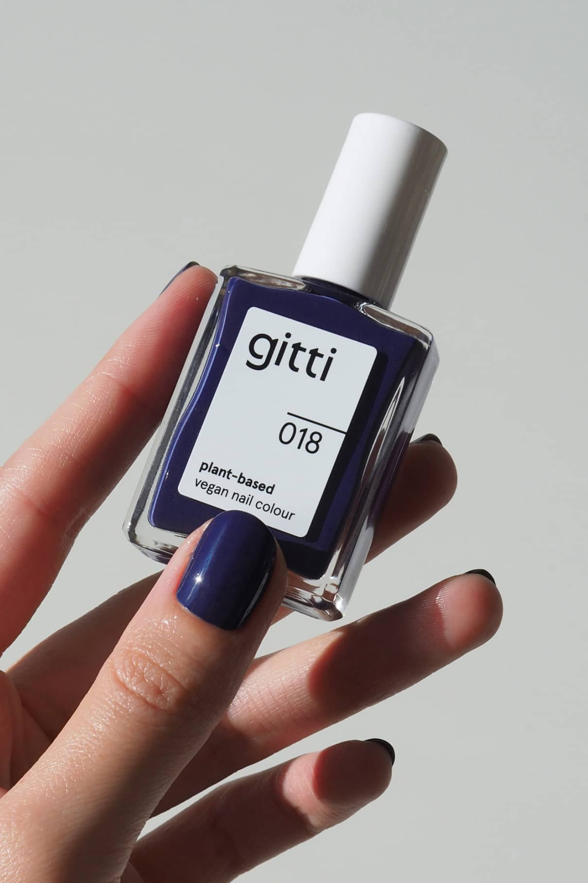 Eine Hand mit lackierten Nägeln hält eine Flasche von gitti no. 018, einer pflanzlichen, veganen Nagelfarbe in einer Mischung aus Tiefblau und Violett.
