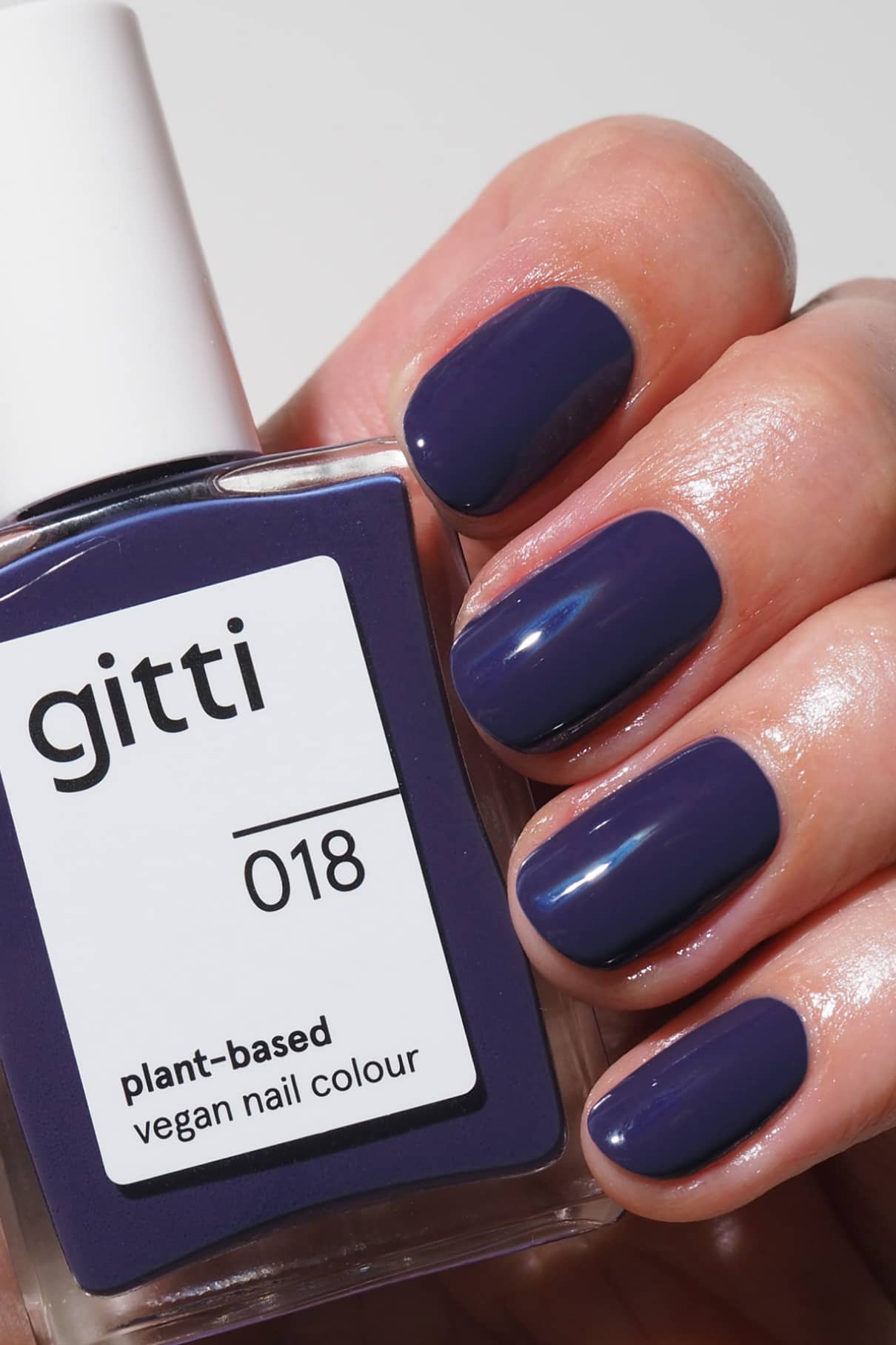 Eine Hand mit sauber manikürten Nägeln, die in einer Mischung aus tiefem Blau und Violett lackiert sind, hält eine Flasche gitti no. 018, einem veganen Nagellack auf Pflanzenbasis, in der gleichen kräftigen Nuance.