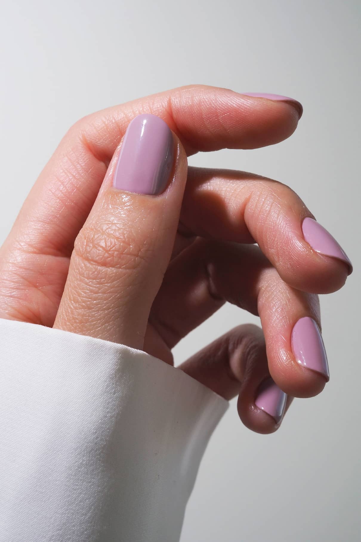Eine Hand mit kurzen, sauber manikürten Nägeln, lackiert in gitti no. 021, einem pudrigen Fliederton, vor einem neutralen Hintergrund, auf dem ein Teil eines weißen Ärmels zu sehen ist.