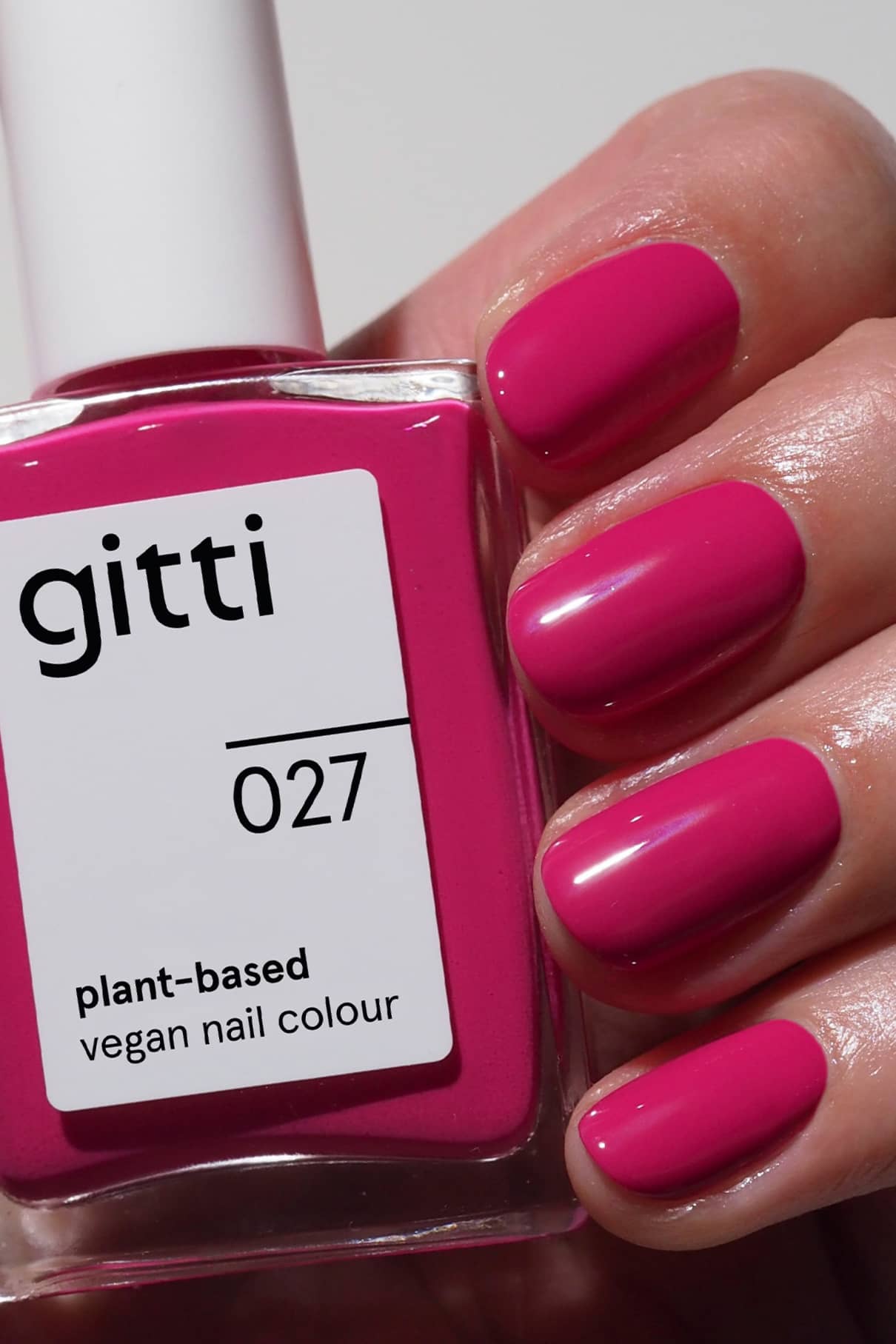 Eine Hand mit kurzen, in leuchtendem Fuchsia-Pink lackierten Nägeln hält eine Flasche gitti no. 027, einem pflanzlichen veganen Nagellack im gleichen Farbton.