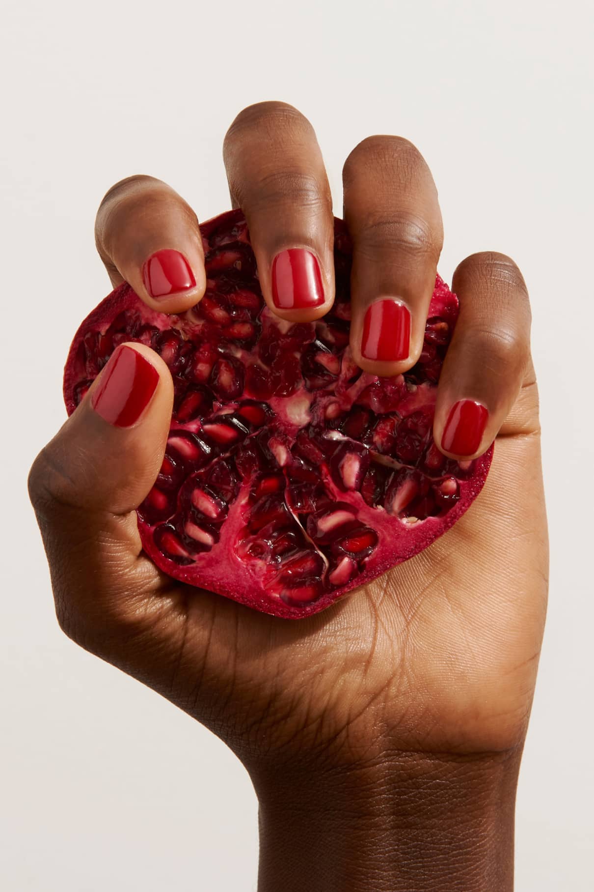 Eine Hand mit Nägeln, die mit Gitti Nr. 102 hält einen halben Granatapfel, dessen Kerne vor einem schlichten hellen Hintergrund glitzern.
