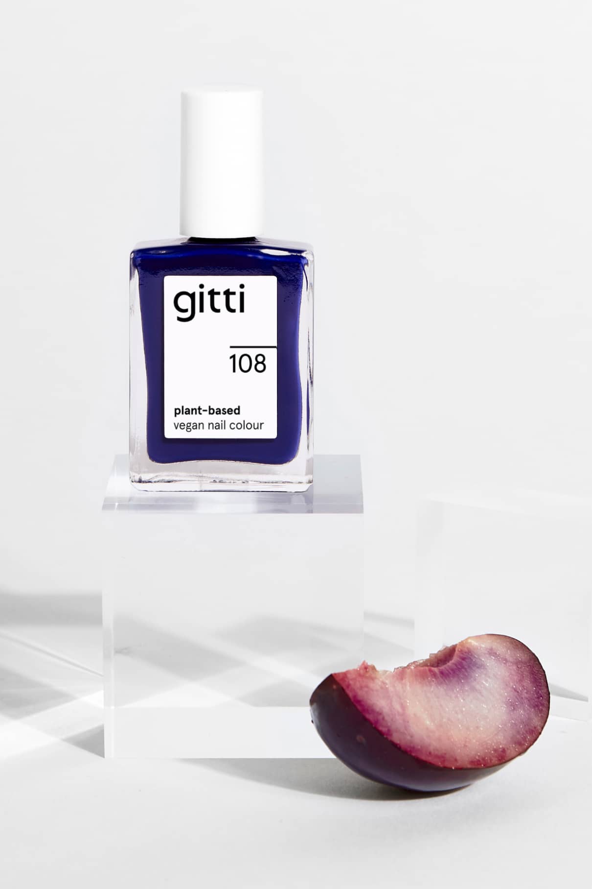 Eine Flasche gitti no. 108 pflanzliche vegane Nagelfarbe in einem intensiven Blauton ist auf einem klaren Acrylblock neben einer aufgeschnittenen Pflaume abgebildet, alles vor einem weißen Hintergrund.