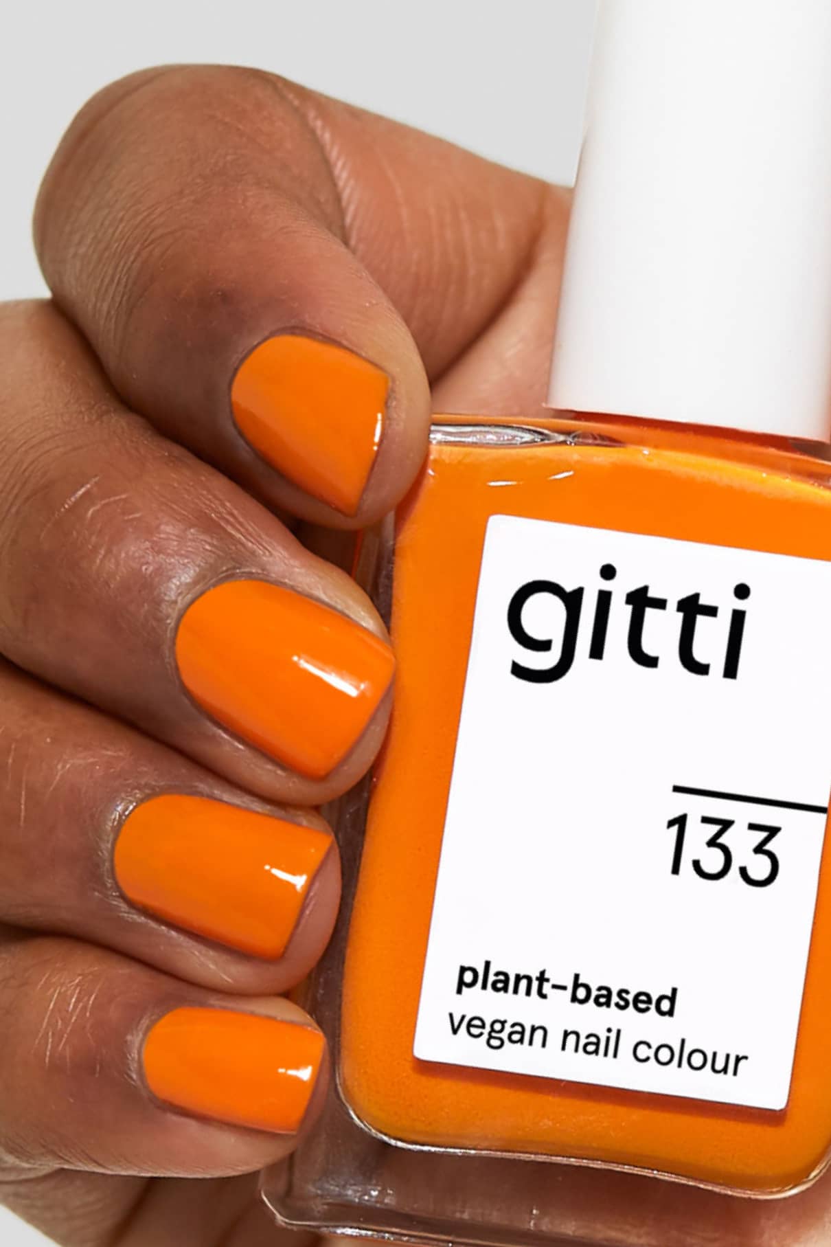 Eine Hand mit orange lackierten Nägeln in warmem Orange hält eine Flasche gitti no. 133 vegane Nagelfarbe auf Pflanzenbasis.