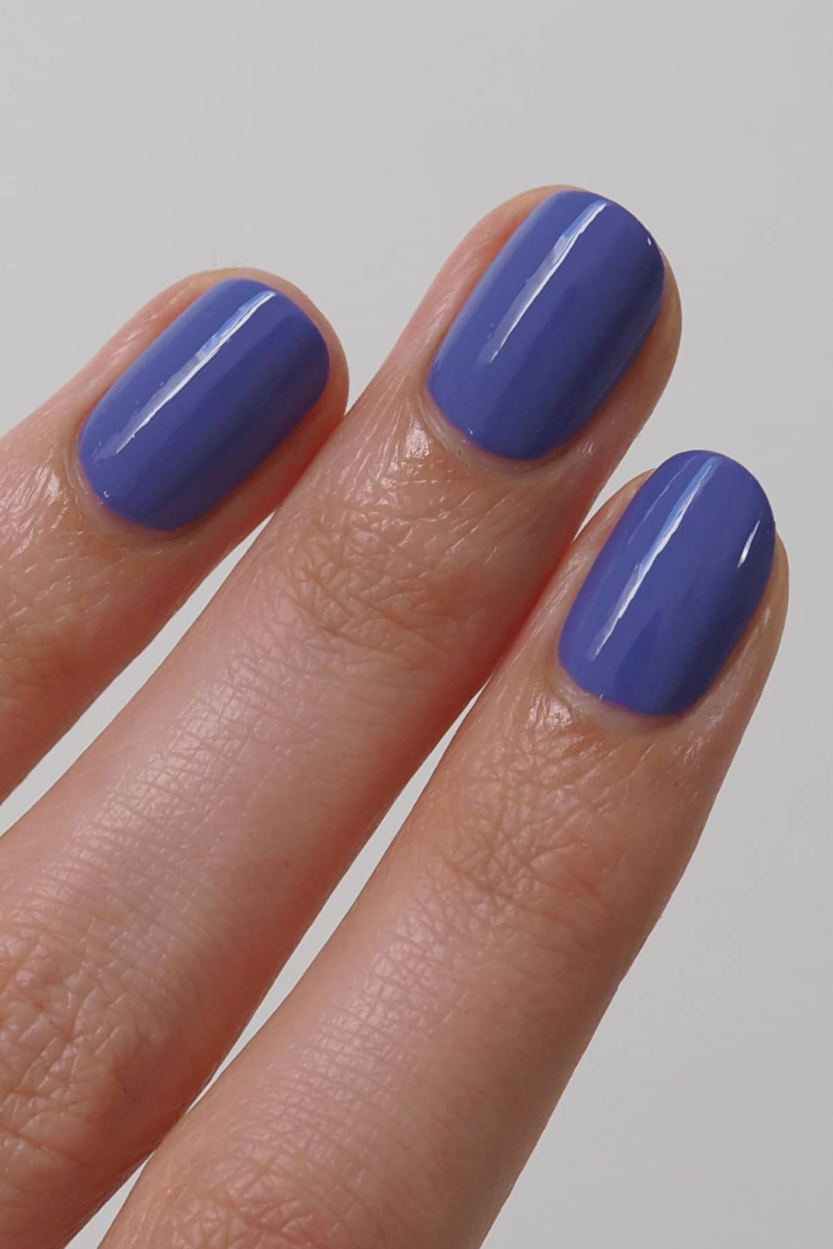 Nahaufnahme von drei Fingern mit kurzen, sauber geformten Nägeln, die in glänzendem Gitti No. 152, einem blaustichigen Violett, inspiriert von Pantone Farbe des Jahres 2022, vor einem unifarbenen Hintergrund.