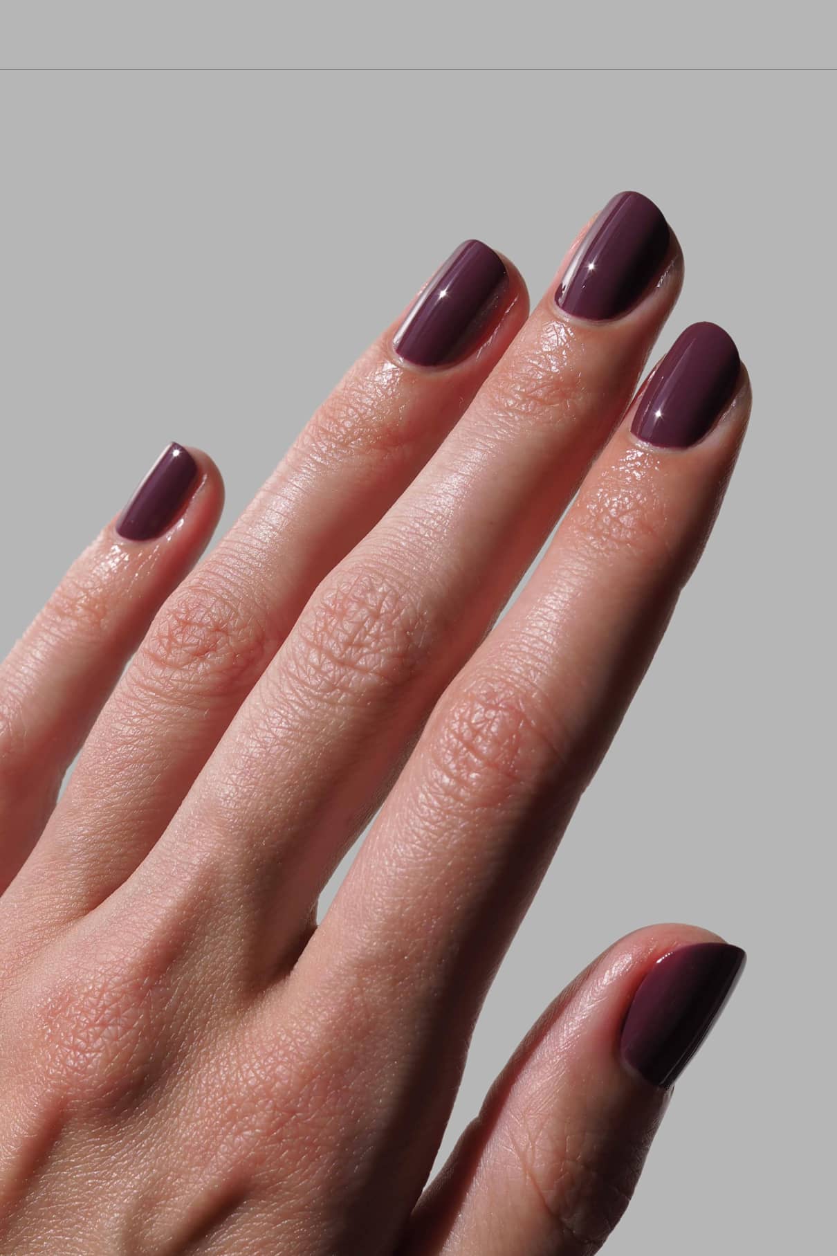 Eine Hand mit kurzen, sauber manikürten Nägeln, lackiert in glänzendem gitti no. 164, einem rötlichen Lila-Lack mit blauen Untertönen, dargestellt vor einem schlichten grauen Hintergrund.