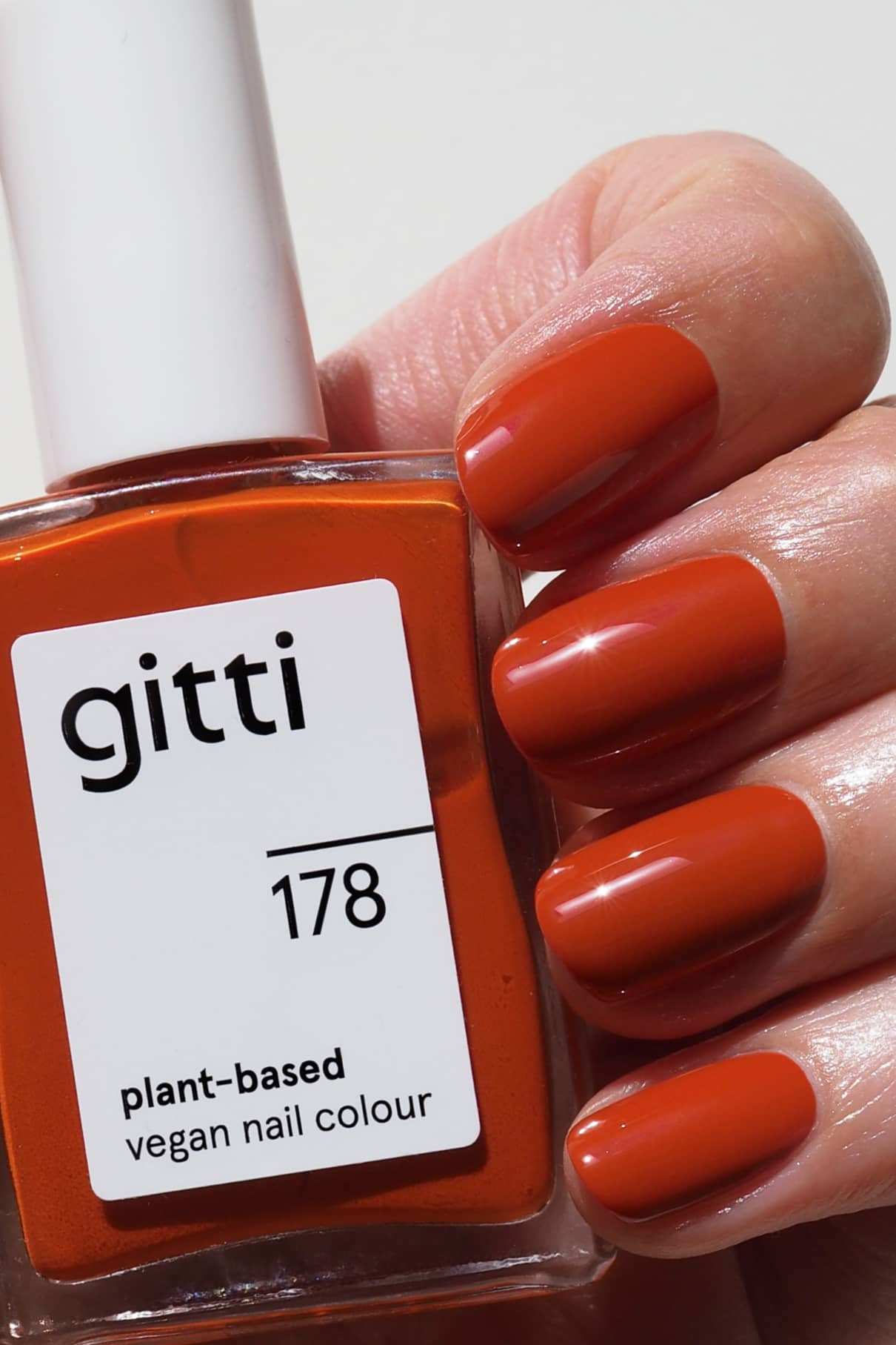 Eine Hand mit sauber manikürten Nägeln in einem warmen, gesättigten Orangerot-Ton hält eine Flasche gitti no. 178 veganen Nagellack auf Pflanzenbasis.