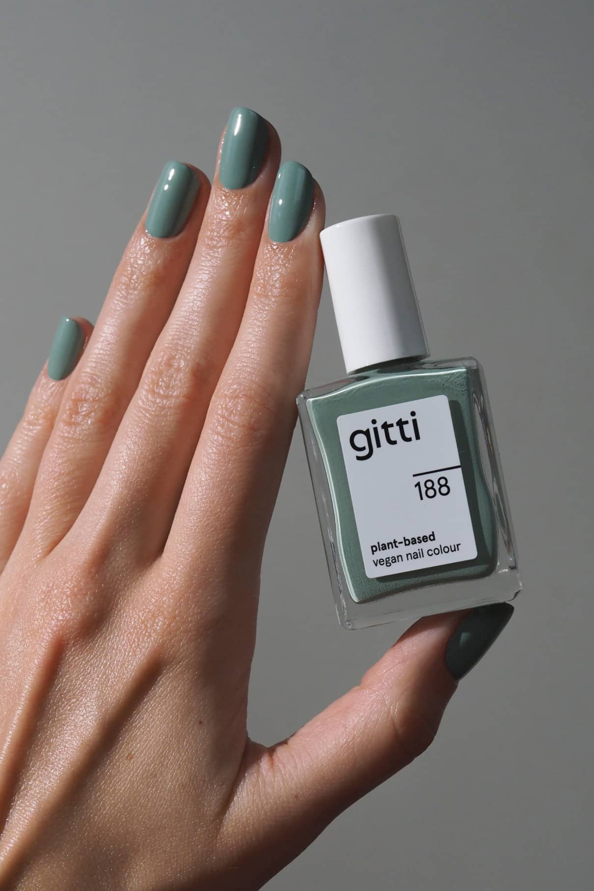 Eine Hand mit grün lackierten Nägeln hält ein Fläschchen mit gitti no. 188 pflanzlicher veganer Nagelfarbe vor einem schlichten grauen Hintergrund.