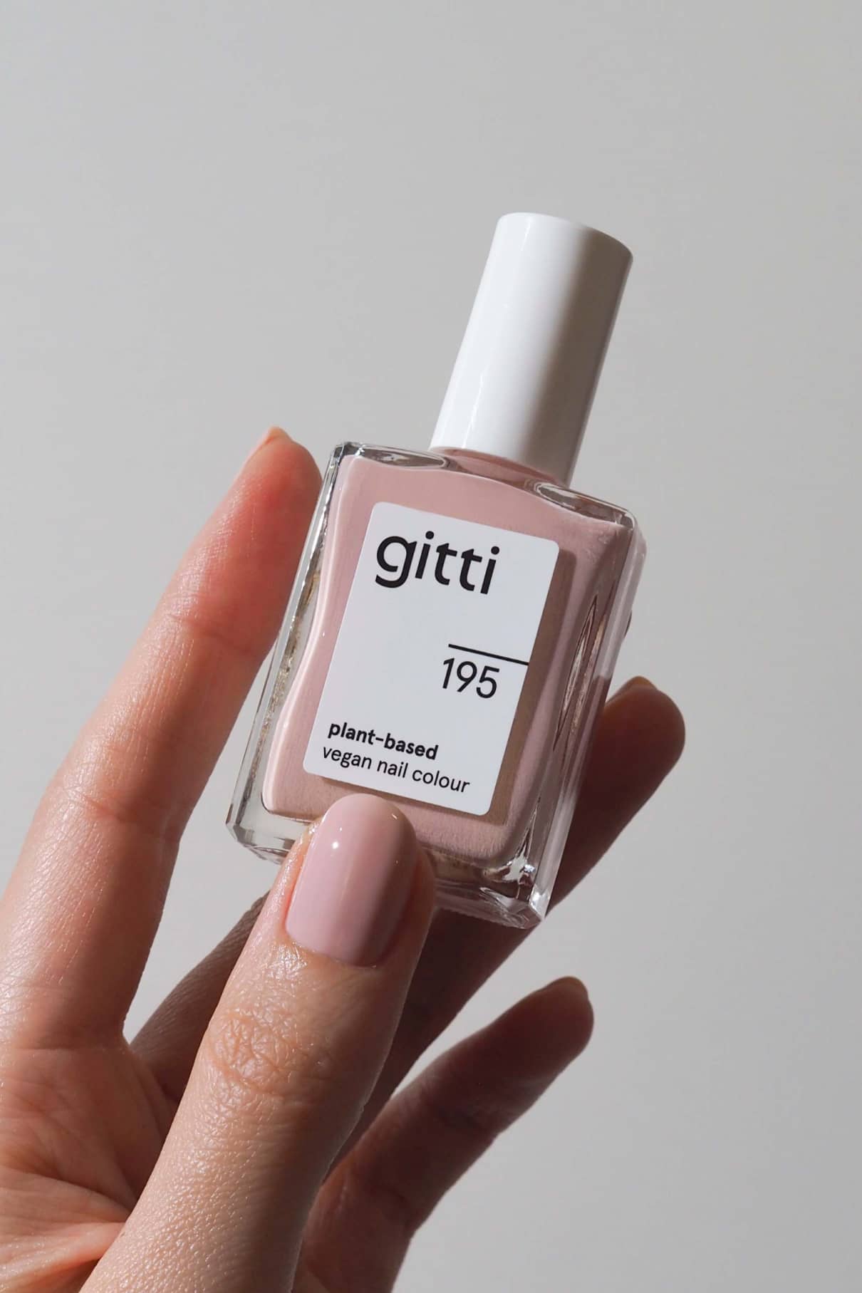 Eine Hand mit blassrosa lackierten Nägeln hält eine Flasche gitti no. 195 pflanzliche vegane Nagelfarbe in pudriges Rosé vor einem schlichten Hintergrund.