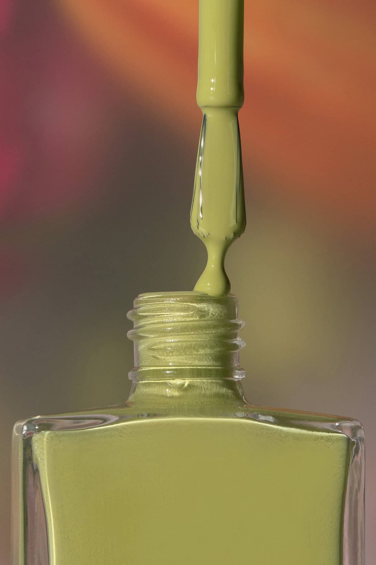 Ein Pinsel trägt den Nagellack gitti no. 196 Nagellack, ein harmonisches Grün, aus einer quadratischen Glasflasche vor einem unscharfen mehrfarbigen Hintergrund.