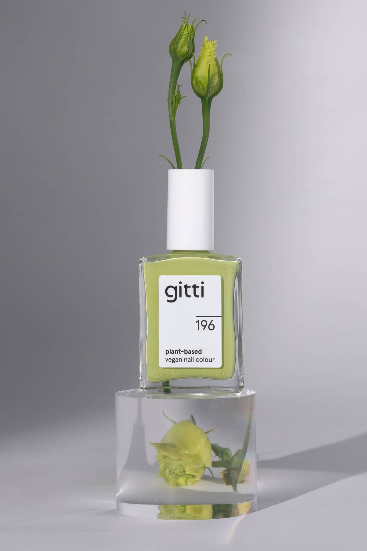 Eine Flasche gitti no. 196 pflanzliche vegane Nagelfarbe, Farbton harmonisches Grün, mit grünen Blütenknospen in der Kappe sitzt auf einem durchsichtigen Block mit einer Blume im Inneren.