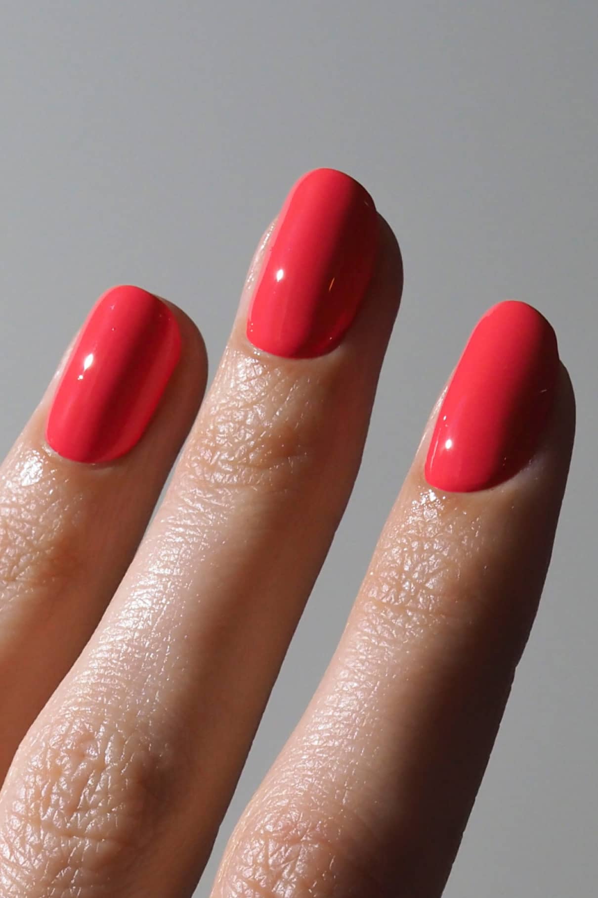 Nahaufnahme von drei Fingern mit glänzendem, hellrosa Nagellack - Gitti Nr. 199, eine leuchtende Mischung aus Rosa und Rot, vor einem schlichten grauen Hintergrund.