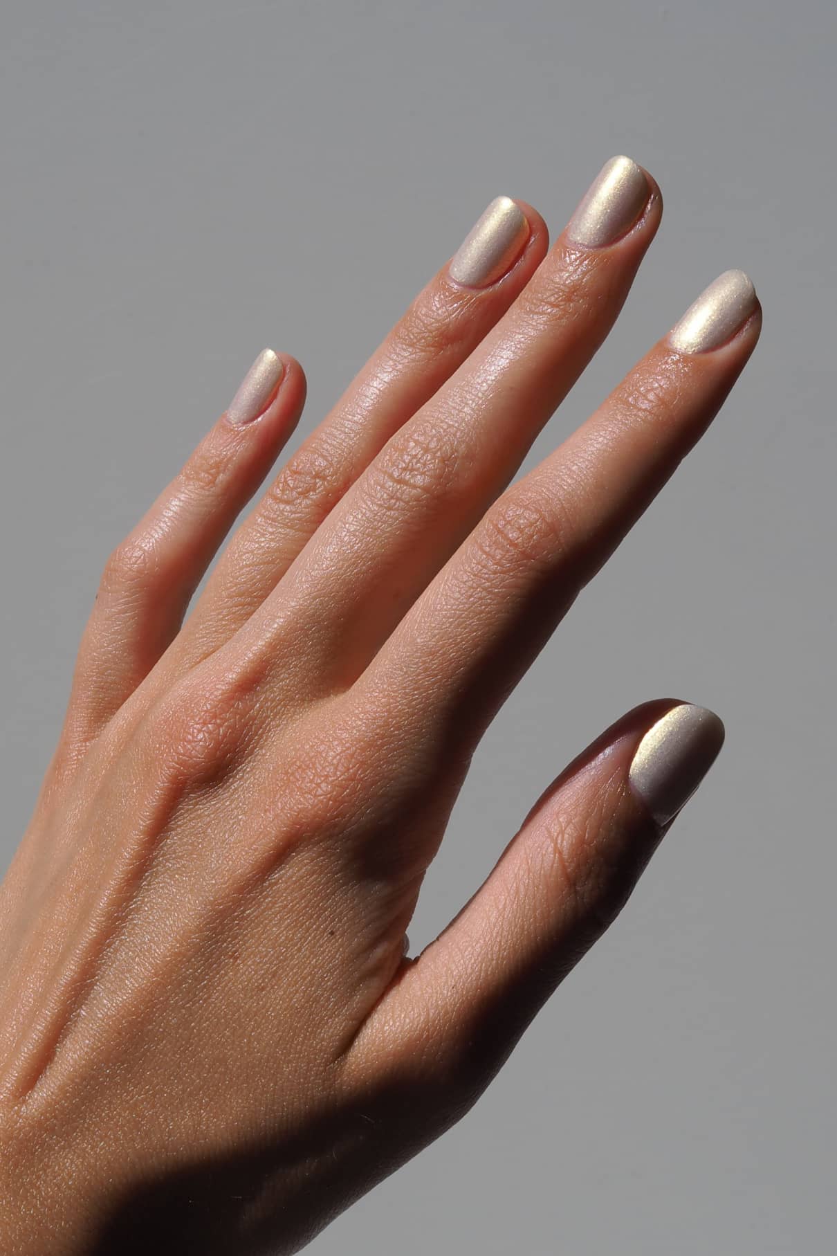 Eine Hand mit sauber manikürten Nägeln, lackiert mit gitti no. 207, einem metallischen gold-bläulichen Lack mit irisierendem Schimmer, vor einem schlichten grauen Hintergrund.