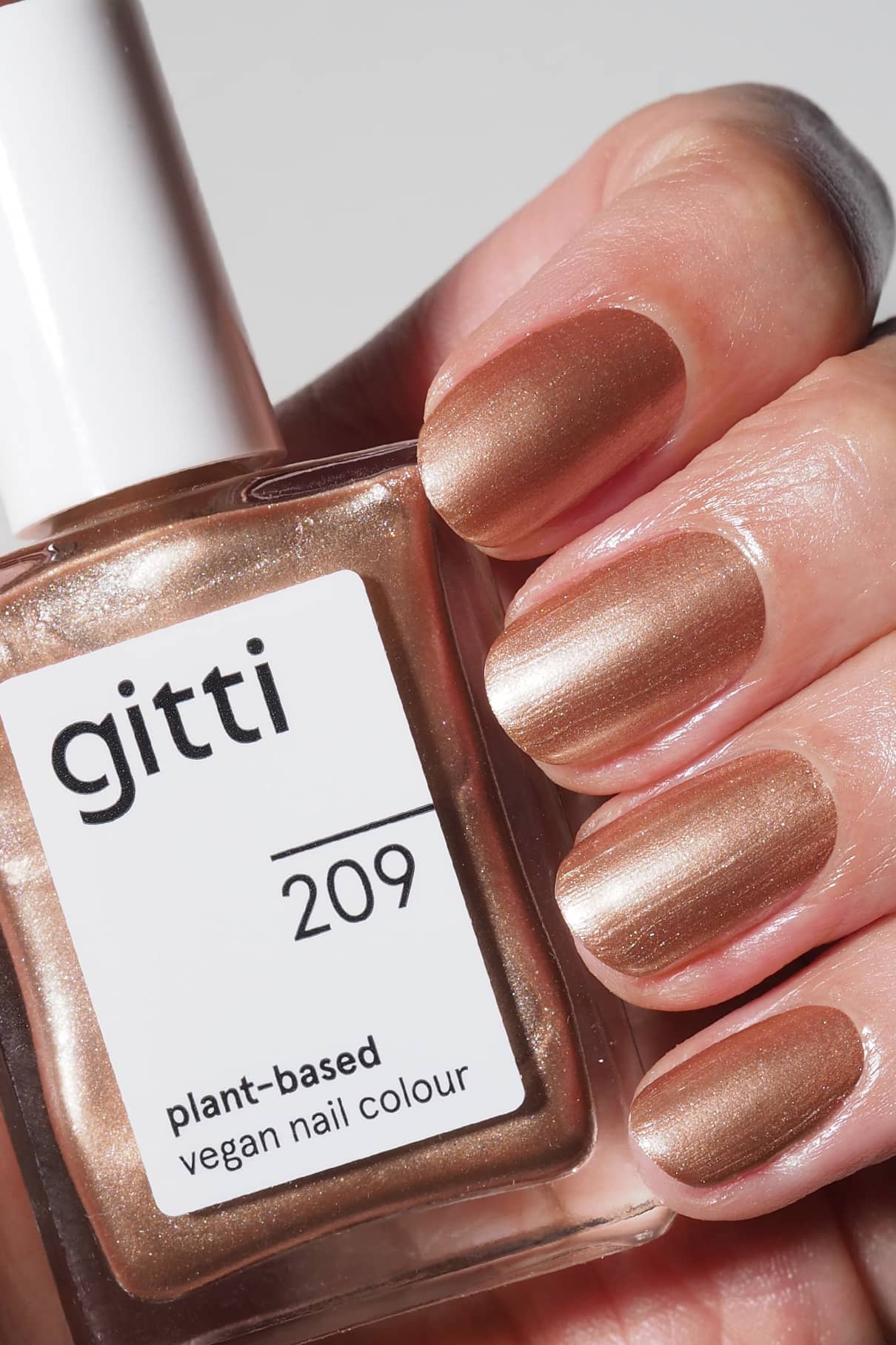 Eine Hand mit kurzen Nägeln, die in einem satten Bronzeton-Metallic-Ton lackiert sind, hält ein Fläschchen mit gitti no. 209 pflanzlicher, veganer Nagellack.