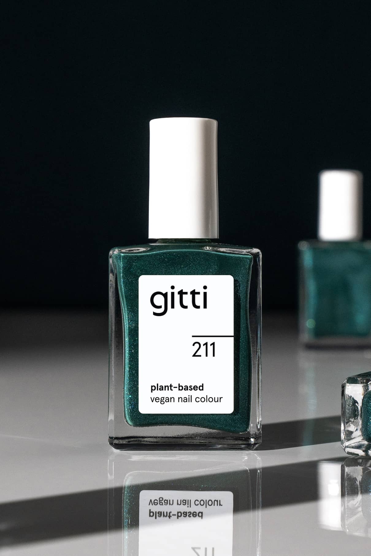 Eine Flasche gitti no. 211 pflanzliche vegane Nagelfarbe in Teal mit silbrig-blauem Schimmer und weißer Kappe steht auf einer spiegelnden Fläche, im Hintergrund ist eine weitere Nagellackflasche zu sehen.