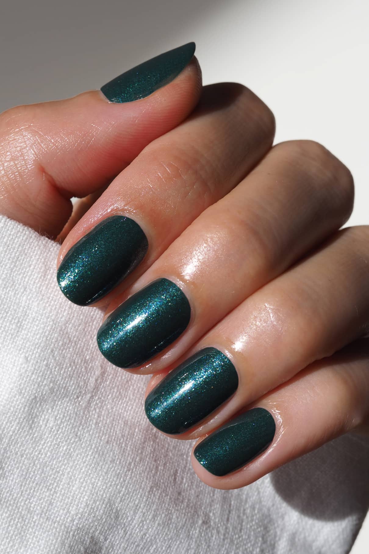 Nahaufnahme einer Hand mit kurzen, gepflegten Nägeln, die in gitti no. 211, einem glitzernden Dunkelgrün mit silberblauem Schimmer, die auf weißem Stoff ruht.