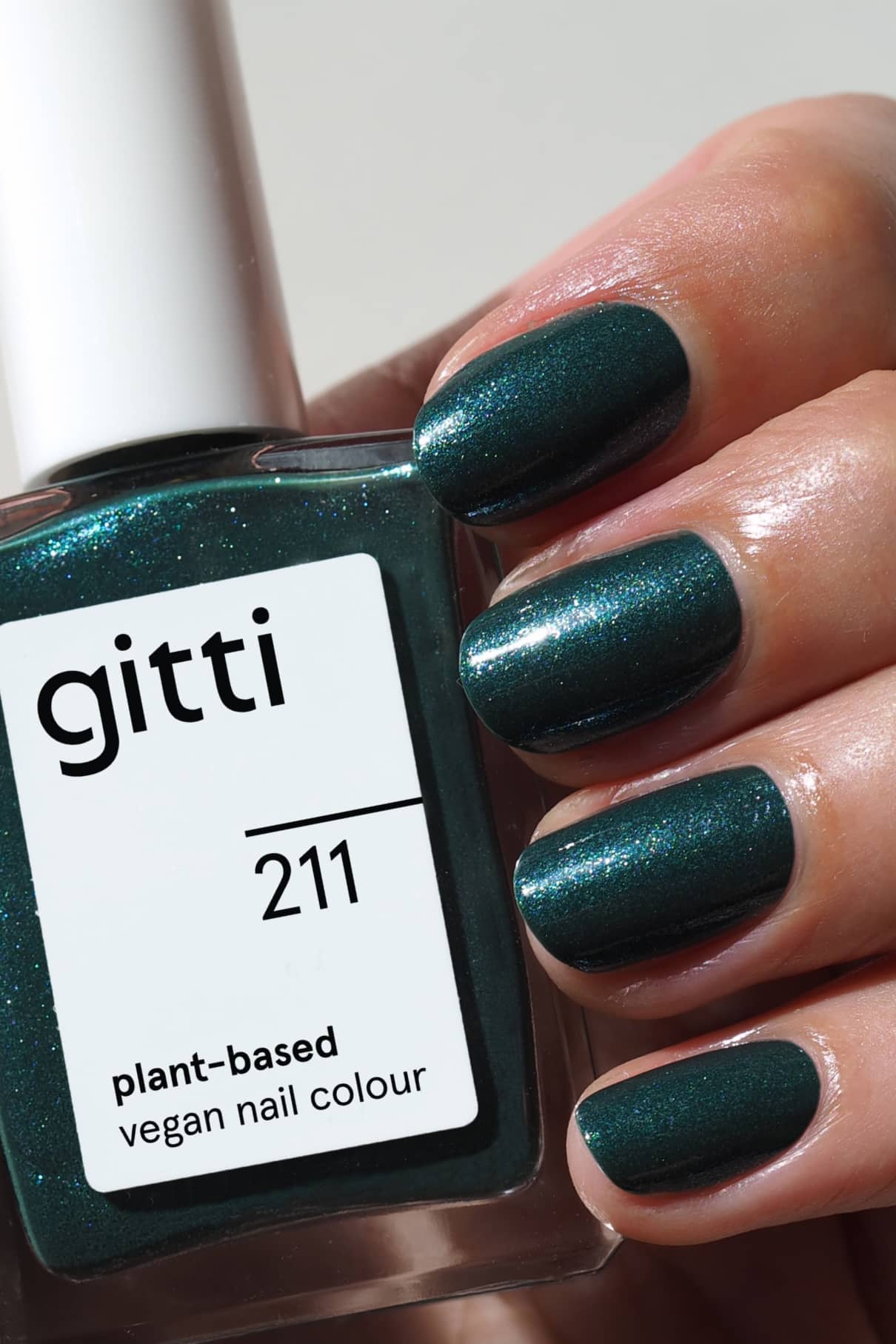 Eine Hand mit glitzerndem Dunkelgrün, schimmerndem Nagellack hält eine Flasche mit der Aufschrift gitti no. 211 pflanzlich-vegane Nagelfarbe.